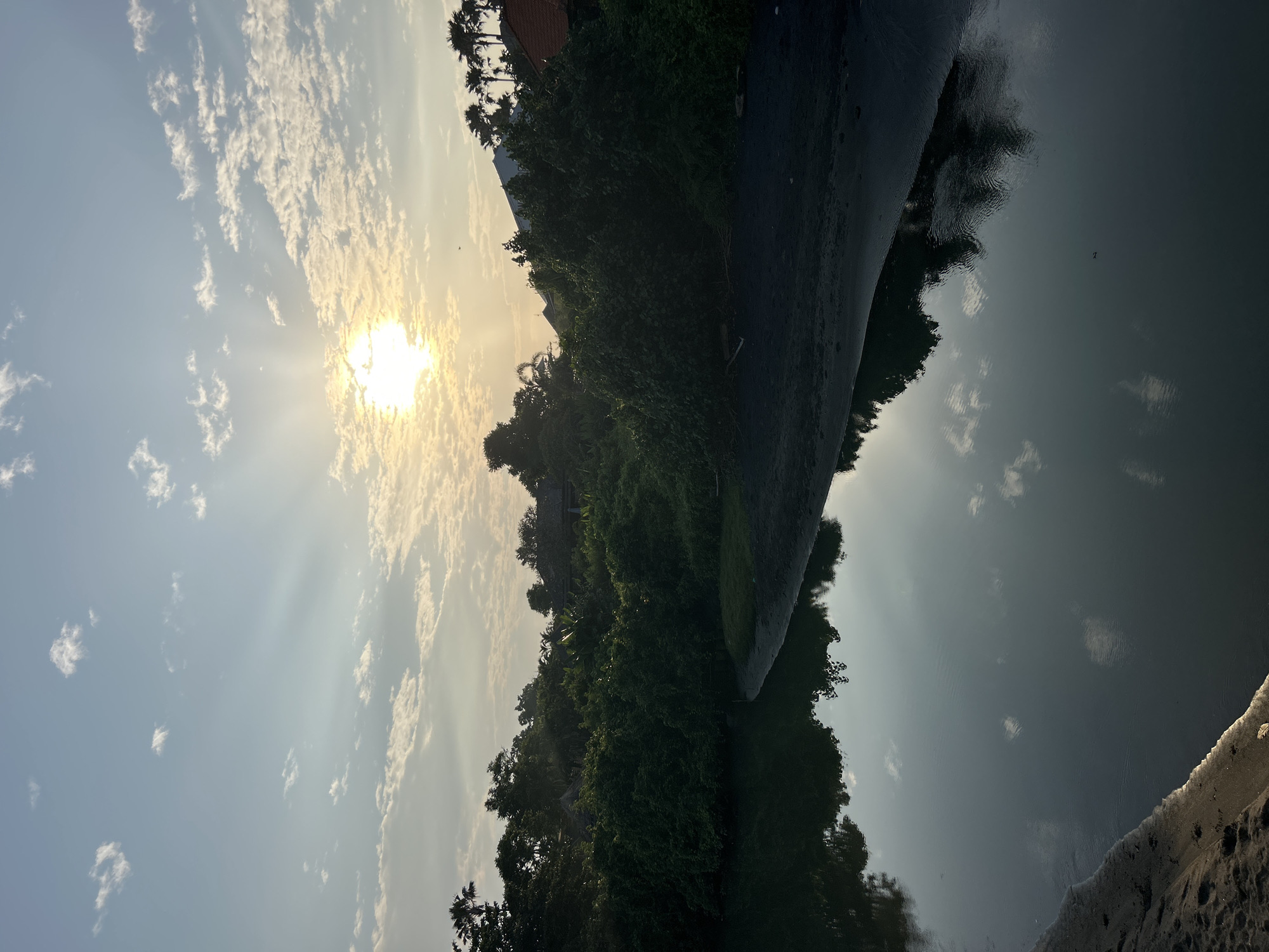 This vertical, outdoor, eye-level shot captures a serene water scene at sunset in Mengwi, Indonesia. The foreground features dark sand and the calm, dark water of a river or bay, reflecting the sky and surrounding trees. The middle ground is dominated by lush, dense green foliage, creating a thick wall of vegetation that lines the water's edge. Peeking through the trees are glimpses of buildings with terracotta roofs and silhouettes of palm trees against the sky. The sky above is a pale blue, transitioning to a soft yellow near the horizon. Sunlight beams dramatically through scattered, wispy white clouds, with the bright sun partially obscured by some of these clouds.