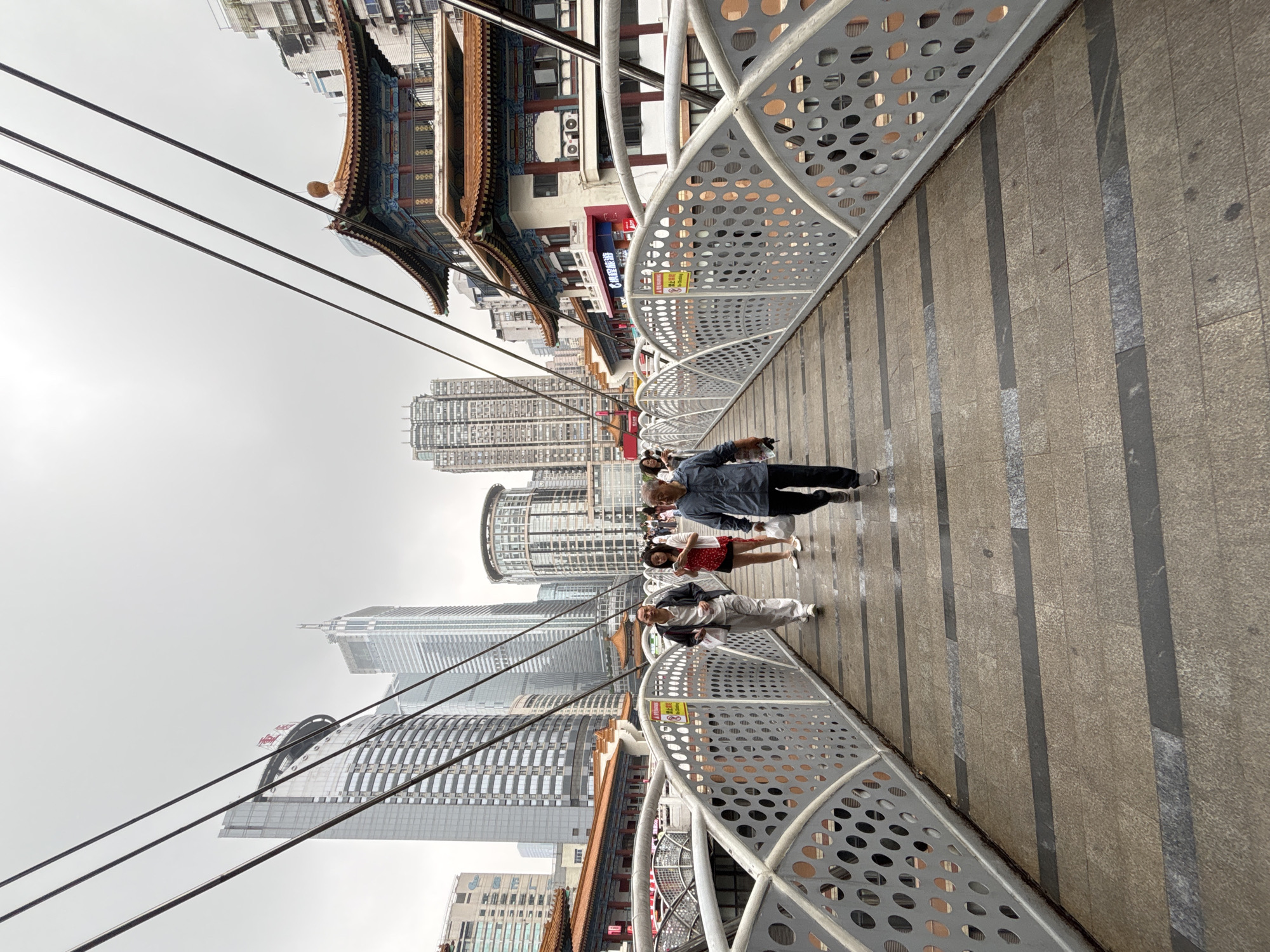 A slightly upward-angled, rotated shot captures a modern bridge in Yuzhong District, China, against a backdrop of skyscrapers and traditional Chinese architecture.
Several people are walking on the bridge. The bridge has a gray metal railing with circular cutouts. Wires from above stretch into the sky.
Traditional Chinese buildings with tiered roofs and decorative eaves are visible on the left, contrasting with modern high-rise buildings in the background. The sky is overcast. The bridge's surface is paved with rectangular tiles.