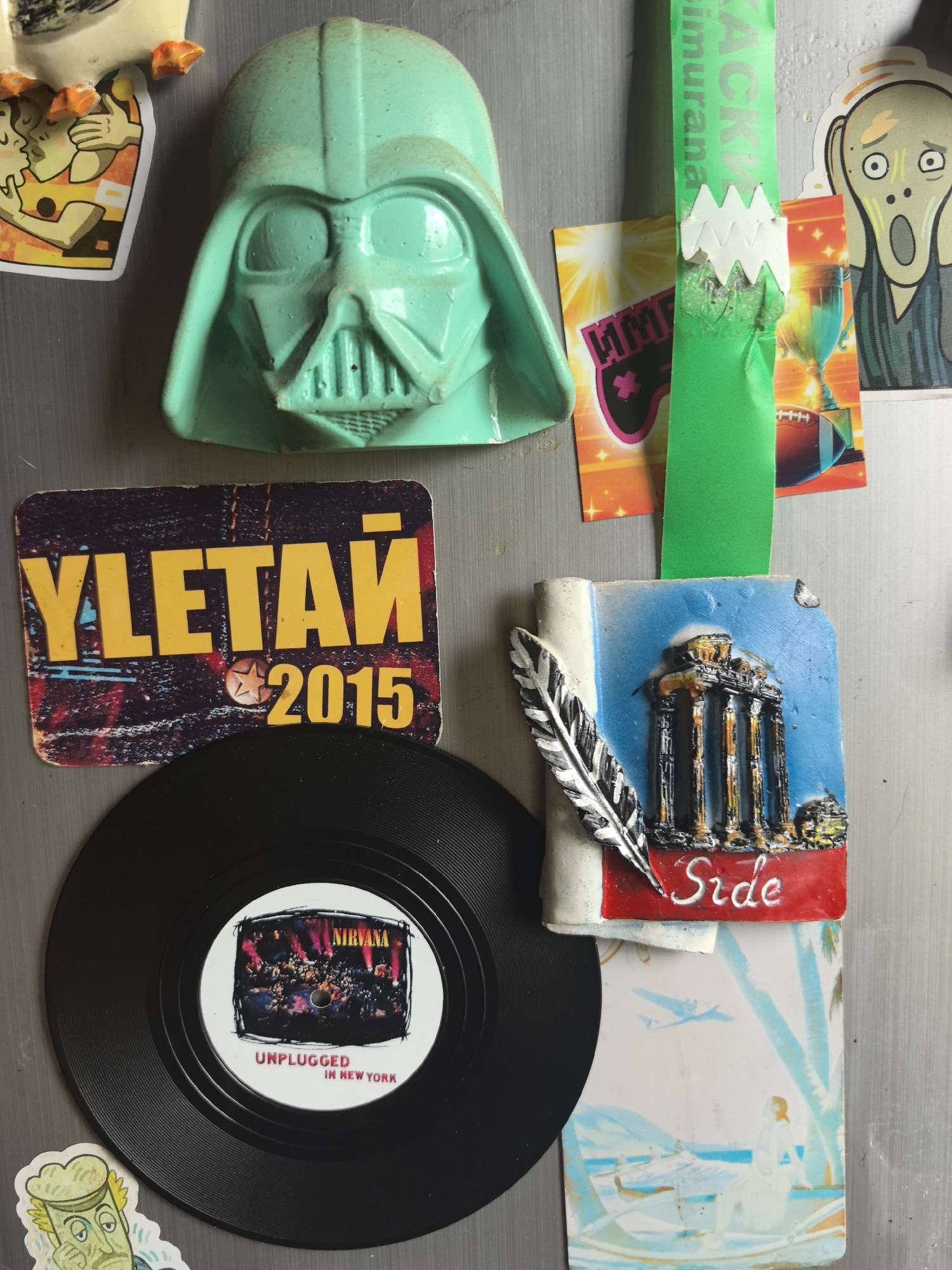 The image is a close-up shot of a refrigerator door covered with various magnets and stickers.
The magnets include: a mint-green Darth Vader mask; a rectangular magnet that reads "УЛЕТАЙ 2015", which translates to "Fly Away 2015"; a miniature black vinyl record with a label indicating "Nirvana Unplugged in New York"; and a rectangular magnet that features a depiction of a Greek building, a feather, and the word "Side".
Stickers feature: a cartoon image of a kissing couple, a piece of branded packaging featuring an image of a football and trophy, a "Scream" inspired image, a cartoon with a beer and glass of beer, and a landscape scene of Noviy, Russia.