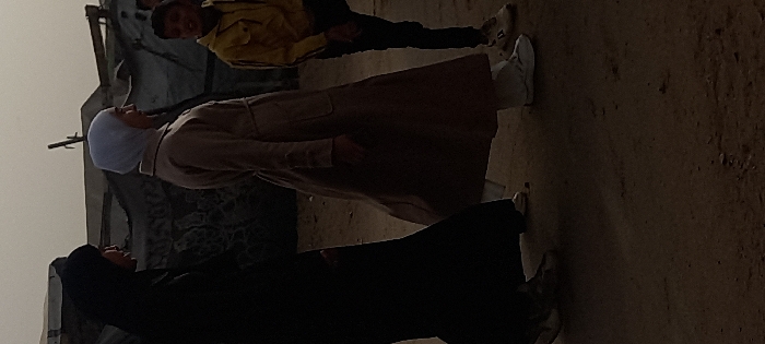 The image depicts an outdoor scene featuring three individuals on an unpaved, dusty ground. The overall lighting suggests it is daytime, possibly overcast or hazy, as there are no harsh shadows.
In the foreground, on the left side, stands a person wearing a long black garment, likely an abaya, and a black head covering that conceals their face, possibly a niqab. They are standing with their body angled towards the right, looking slightly upwards towards the person in the middle. They are wearing light-colored footwear.
In the center of the frame, another person is standing, dressed in a light brown or beige long coat or dress, reminiscent of an abaya, and a light blue or white head covering (hijab). This individual is facing slightly towards the left side of the frame (relative to their upright position), with their face visible in profile. They are wearing white sneakers.
Further in the background, towards the right, a young person, possibly a child or teenager, is visible. They are wearing a bright yellow long-sleeved top and dark trousers. This person is looking generally towards the viewer, with a slight smile on their face. Their hands are down at their sides, and they are wearing dark shoes.
The background consists of rough, possibly dilapidated structures or walls made of earth or stone, with visible textures and some debris. The sky above is a uniform light grey or tan, blending into the distant structures. The ground is uneven dirt or sand, with small rocks and irregularities. The individuals appear to be standing and observing, possibly engaged in a casual interaction or simply present in the outdoor environment. There is no visible text in the image.