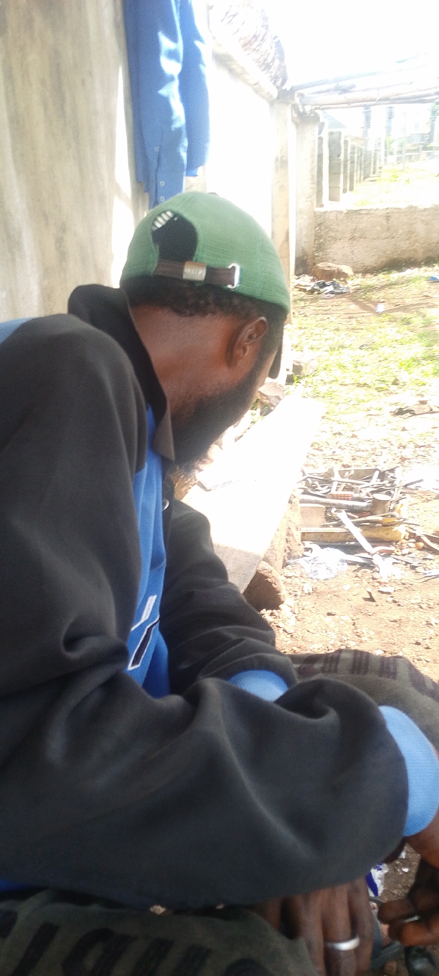 A man with a beard and wearing a green cap, blue shirt, and black jacket is sitting outdoors. He has a ring on his finger. In the background, there is building construction, scattered debris, and green vegetation. The location appears to be a yard or construction site in Lokoja, Nigeria.
