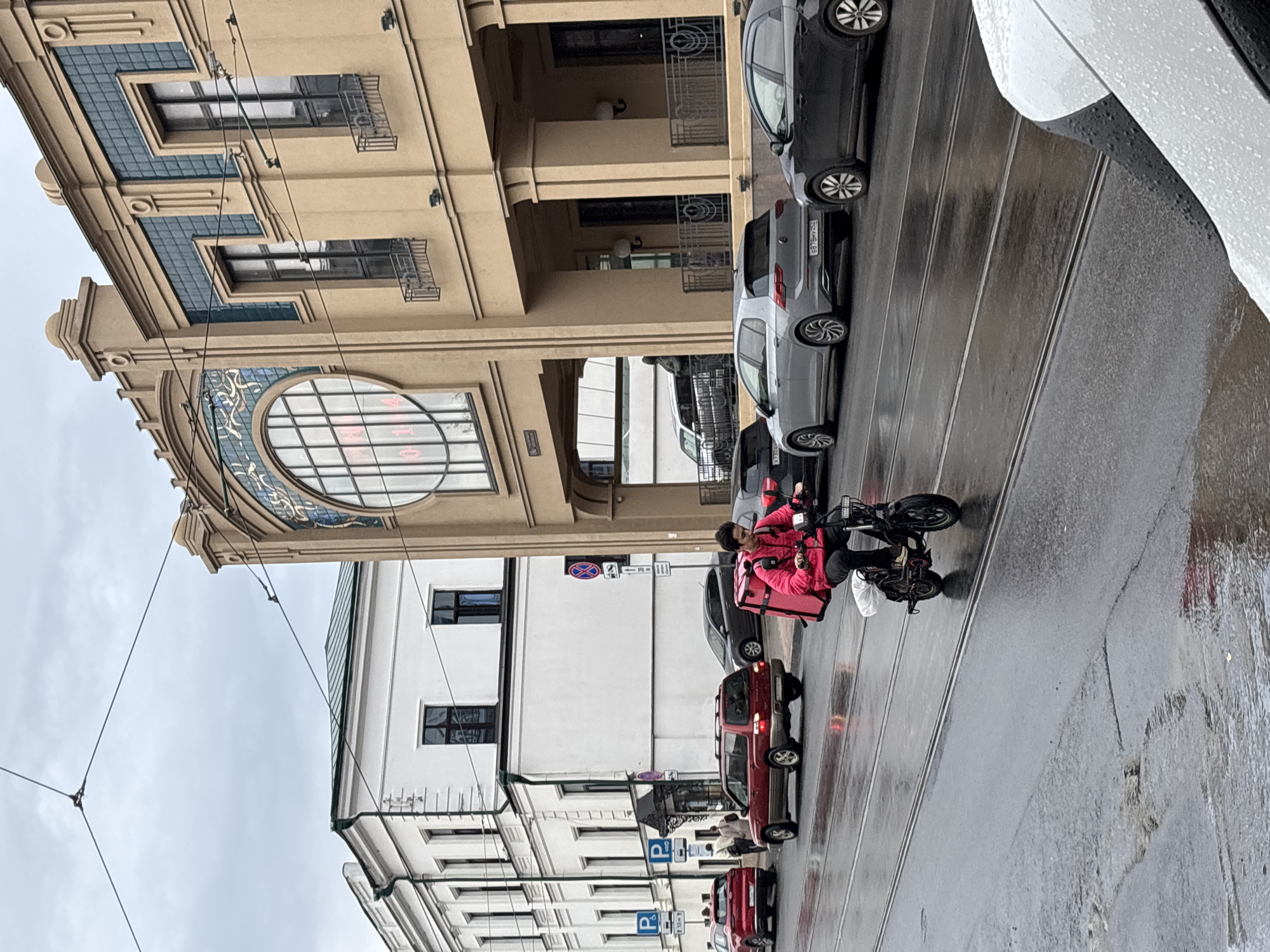 The image shows a street scene in Nizhny Novgorod, Russia. A person wearing a bright pink jacket and carrying a large pink delivery bag on their back is riding a black electric scooter. The scooter has a white bag attached to the front. The person is on a wet asphalt road with tram tracks. In the background, there are several cars, including a red SUV, a silver hatchback, and a dark sedan. Behind the cars are buildings; one is a beige building with ornate architectural details, including a large circular window with intricate patterns. Adjacent to it is a white building with dark windows. The sky is overcast.