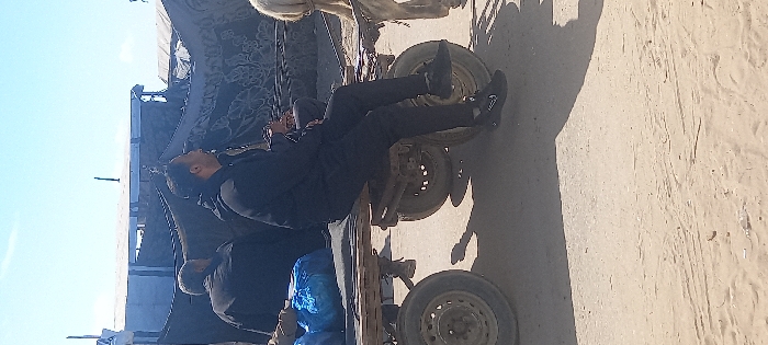 Two men in dark clothing are working on the underside of a horse-drawn carriage. The carriage has four wheels and is parked on a sandy surface. One man is lying on his back beneath the carriage, apparently making repairs. Blue tarp or material is visible near the carriage. The area appears to be an outdoor location, possibly a roadside or rural setting.