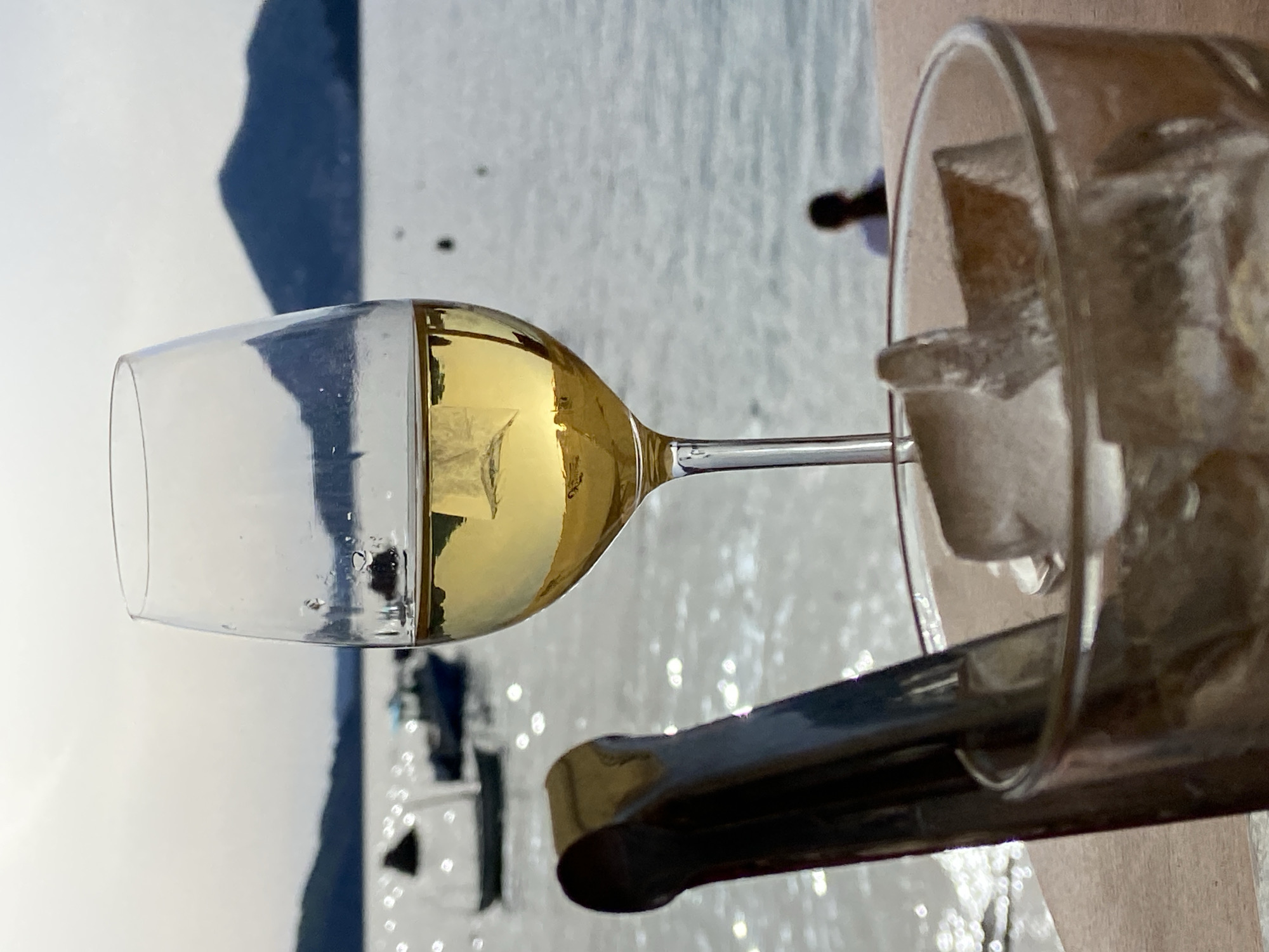 The image depicts a glass of white wine beside a glass filled with ice. Ice tongs rest on the edge of the ice-filled glass. The background shows a body of water with boats and a hilly shoreline, identifiable as Pržno, Montenegro. The photo is taken outdoors, likely on a table. There are no people visible directly, but the placement of the drinks suggests human presence.