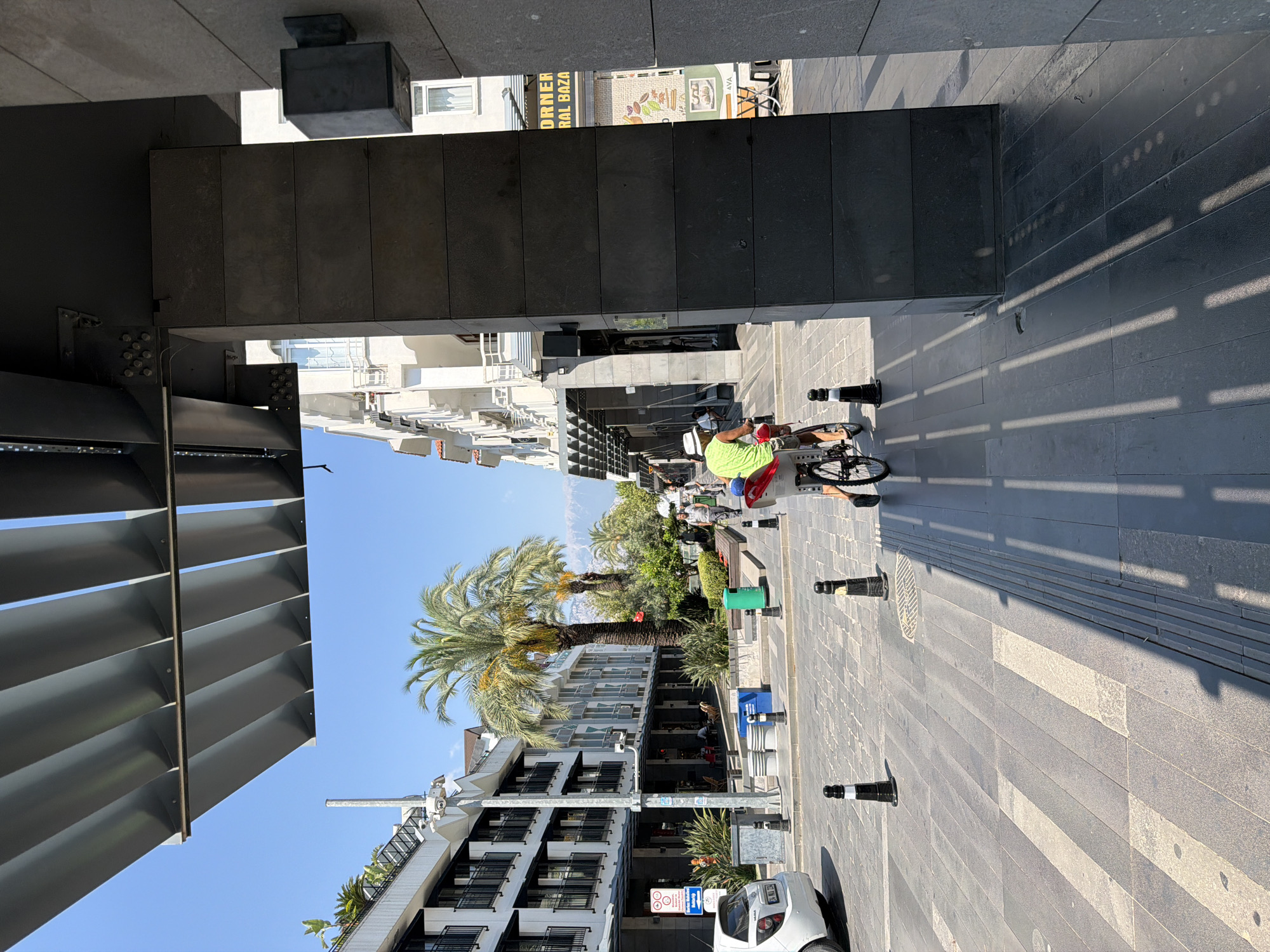 This image depicts a sunny day on a pedestrian street in Kemer, Turkey, likely in an area with shops and hotels. The scene is framed by a modern, dark gray building with a slatted overhang in the foreground. To the left, a white car is parked, and further down the street, white buildings with balconies and black railings line the walkway. A tall palm tree stands prominently in the mid-ground, with smaller green plants and bushes also present. A person wearing a bright green shirt and a white hat is riding a bicycle away from the camera, their legs visible as they pedal. The street is paved with stone tiles, and shadows from the overhang create a pattern on the ground. In the background, distant mountains are visible under a clear blue sky. A sign that reads "CORNER RAL BAZA" is visible on the right side of the frame, suggesting a market or shop.