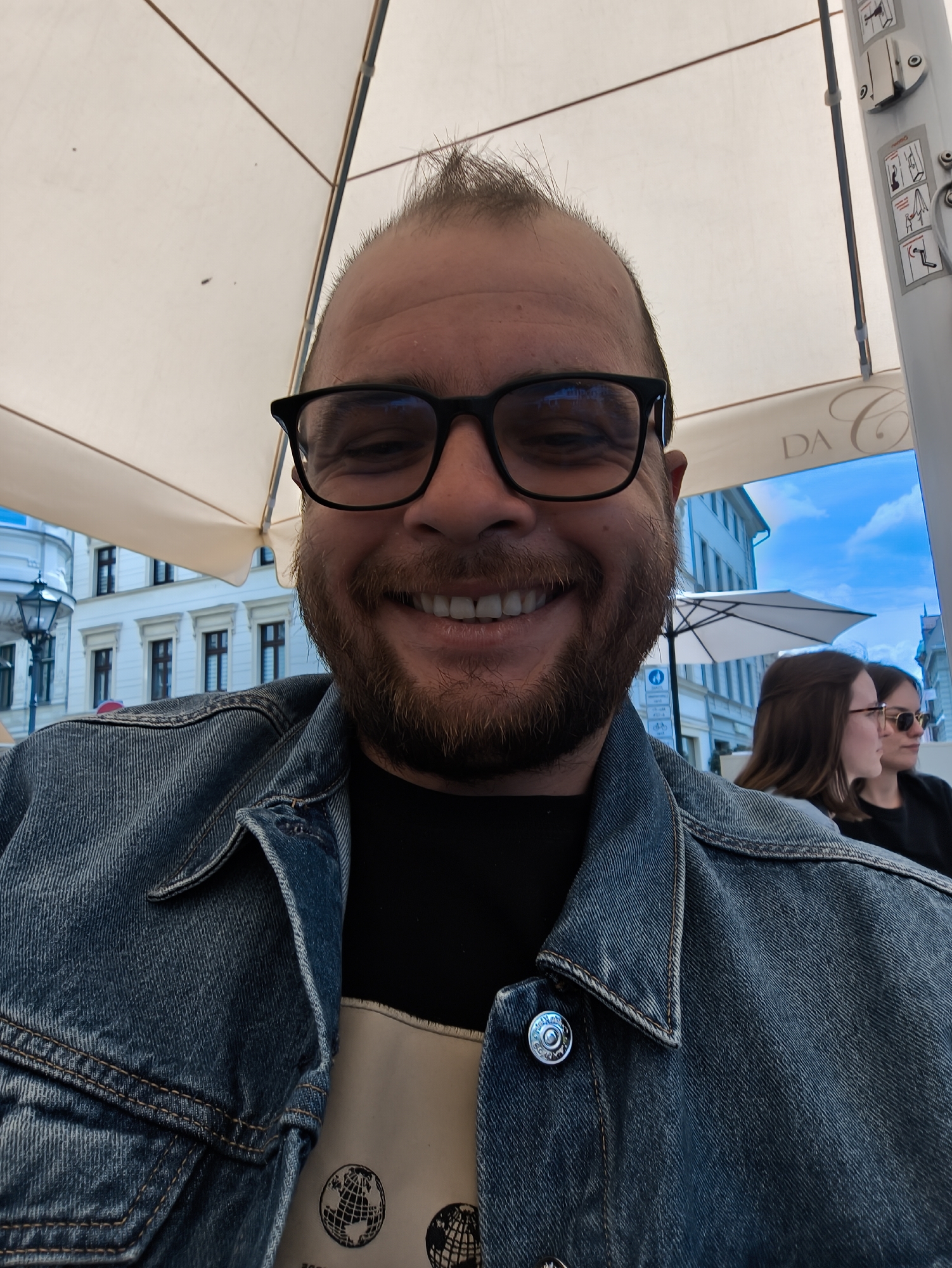 A person is sitting outdoors, smiling at the camera. The person has short hair and a beard and is wearing glasses and a denim jacket. In the background, there are buildings and two people sitting at a table. The setting appears to be a cafe or outdoor dining area, as there is an umbrella overhead. The cafe appears to be named "DA C".