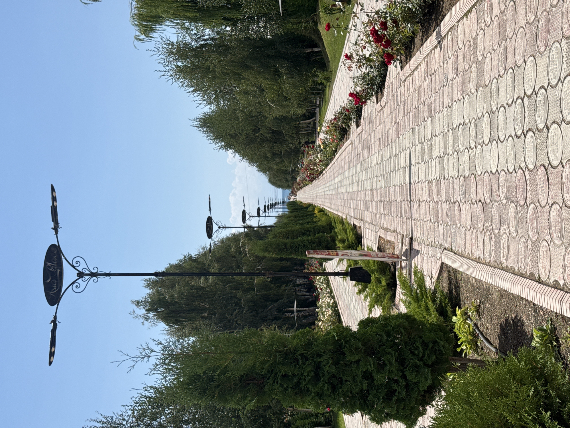 The image captures a paved pathway lined with trees and flowering bushes, extending towards a distant body of water under a clear blue sky. The pathway is constructed of light-colored rectangular tiles with a decorative pattern, bordered by smaller, darker tiles. On the left side of the path, a tall, conical evergreen tree stands prominently, with smaller, bushy evergreens and some low-lying plants visible further down. A signpost is partially visible on the left side of the path.
On the right side of the pathway, a row of rose bushes with red and white blooms adds a splash of color. Both sides of the path are densely lined with trees, creating a tunnel-like effect. Several ornate lampposts with decorative tops and multiple light fixtures are spaced along the pathway, illuminating the scene. The "Asia Palace" is visible on the sign of the lamppost in the foreground.
In the distance, the pathway leads to a hazy horizon where the sky meets what appears to be a body of water, possibly Lake Issyk-Kul, given the location. The overall impression is one of a well-maintained park or promenade, likely a tourist attraction in Sary-Oy, Kyrgyzstan, designed for leisurely strolls and enjoyment of the natural scenery. The scene is peaceful and devoid of overt activity, suggesting a quiet time of day or a less crowded period. The lighting indicates it is daytime, with the sun casting shadows.
