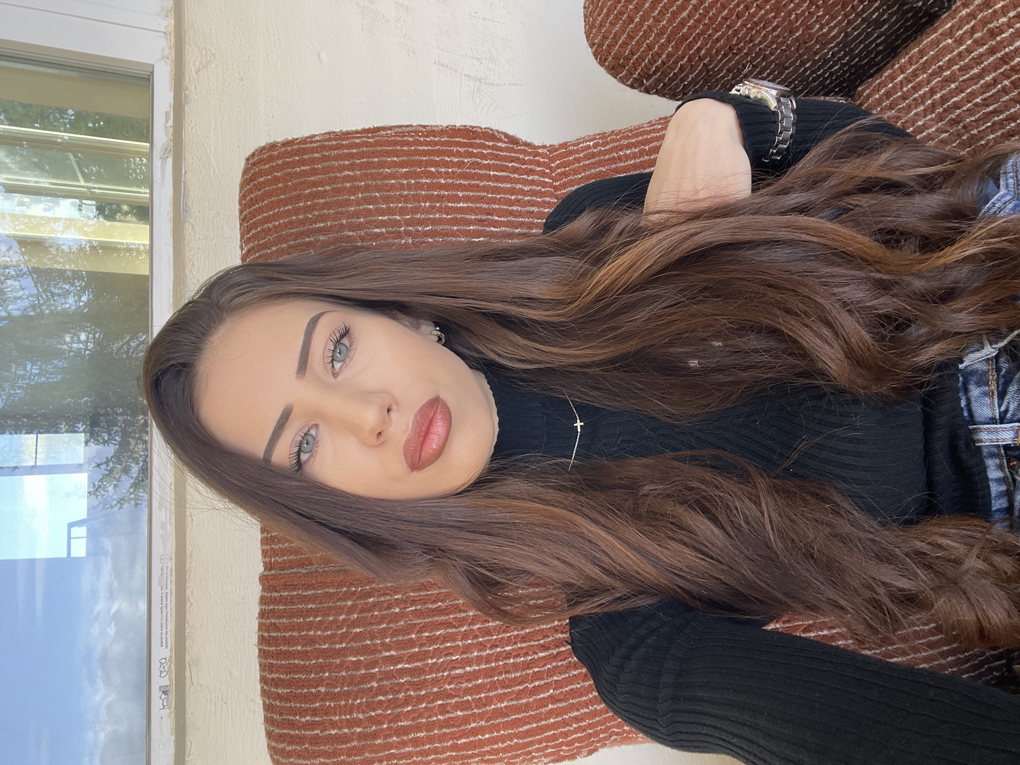 The image is a medium-shot photograph of a young woman with long, brown hair and light-colored eyes. She is reclining on a brown and orange striped fabric surface, likely a sofa or cushions. She wears a black ribbed sweater and a silver watch on her wrist. A silver necklace with a small cross pendant is visible on her chest. Her makeup includes defined eyebrows, light eye makeup, and a neutral lipstick.
To the left of the woman is a window frame with glass panes reflecting the exterior environment, partially obscuring the scene outside.
No specific details indicate the location as Veliko Tarnovo, Bulgaria.