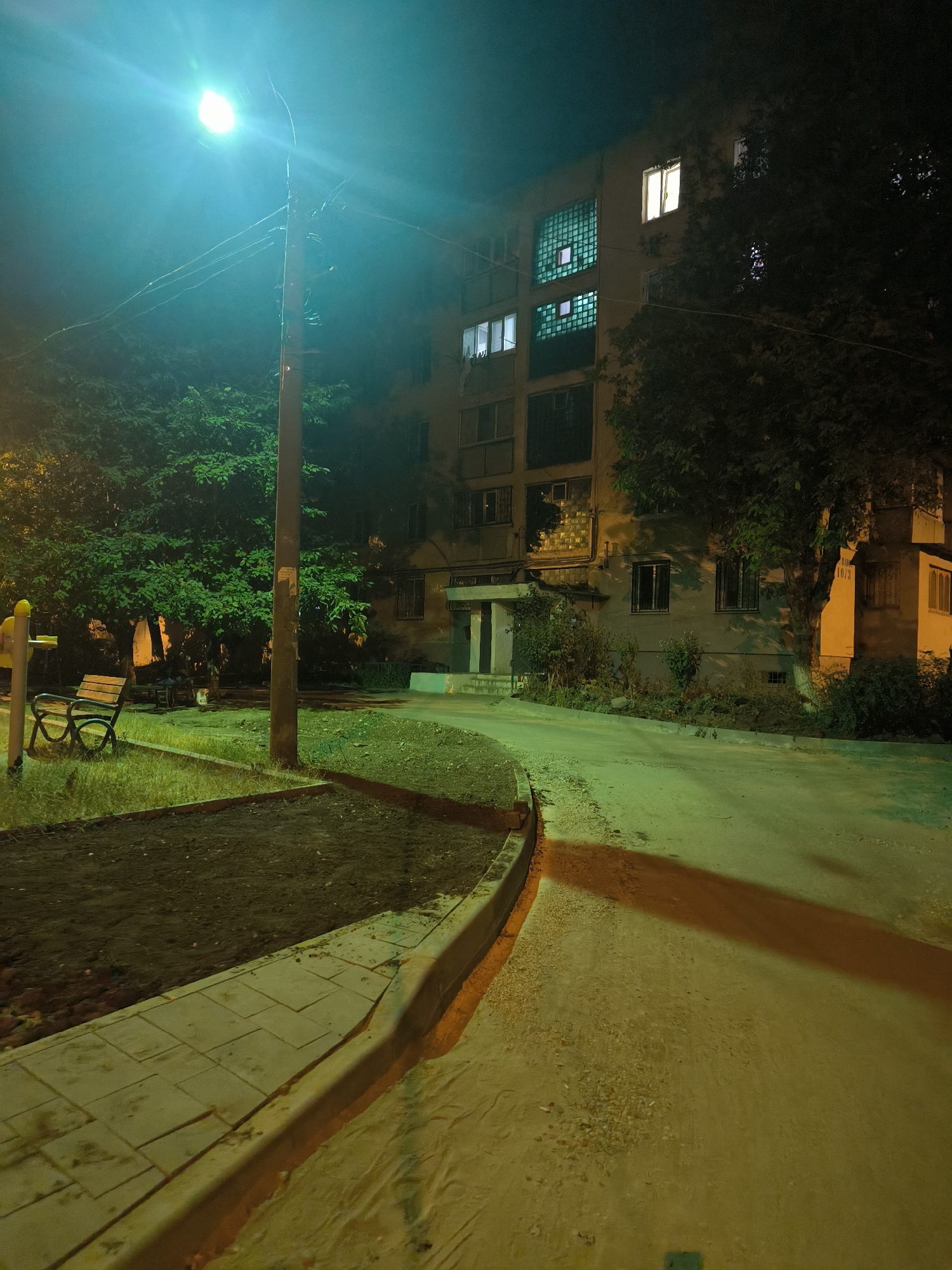 The image depicts a nighttime scene in a residential courtyard in Chișinău, Moldova. A street lamp emits a bright, teal-colored light, casting long shadows. In the foreground, a paved walkway with large square tiles curves to the right, bordering a raised bed of dark soil. A curved concrete curb separates the walkway from a grassy area where a wooden bench with a metal frame sits. To the left of the bench is a yellow playground structure.
Behind the grassy area and bench, a dense cluster of green trees is visible, partially obscuring a multi-story apartment building. The building, typical of Soviet-era architecture, has several lit windows on its upper floors, indicating occupancy. Some windows are fitted with decorative glass blocks. A wooden utility pole stands near the center of the frame, carrying electrical wires. The ground is a mix of asphalt and gravel, with a pronounced orange shadow cast across the asphalt from the street lamp, suggesting it is angled towards the building and walkway. The overall atmosphere is dimly lit and somewhat eerie due to the unusual lighting conditions.