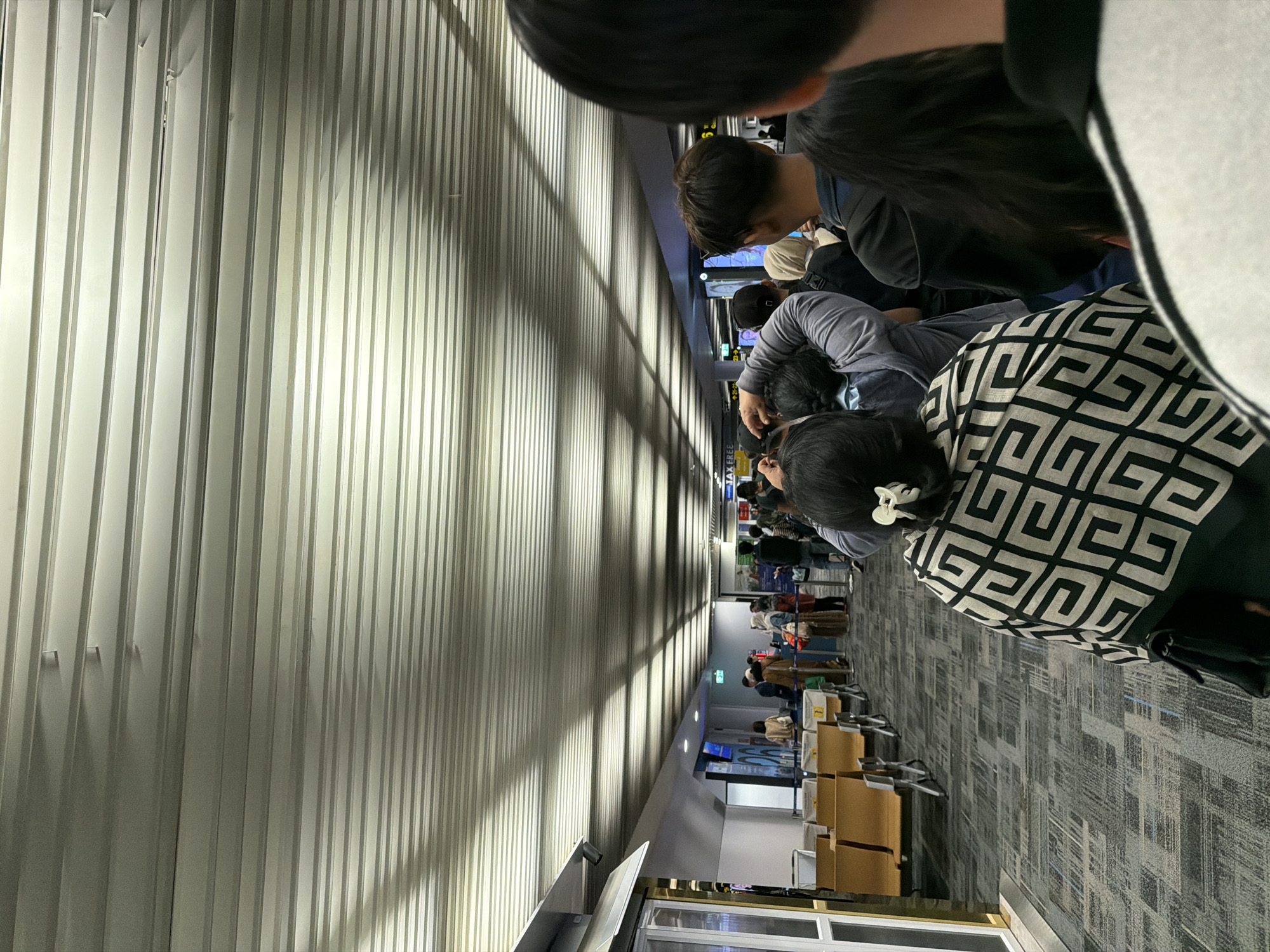The image depicts a queue of people inside an airport terminal, likely at Suvarnabhumi Airport in Bangkok, Thailand, given the context of travel. The ceiling features a distinctive ribbed, metallic design, a common architectural element in modern airports. Fluorescent lighting casts long shadows across the ceiling and down the hallway. In the foreground, the back of a person wearing a patterned shawl is visible. Further down the queue, various individuals can be seen with luggage, suggesting they are either arriving or departing. The environment appears orderly, with seating areas visible to the left and information displays or signs further down the terminal. The overall scene conveys a sense of transit and waiting, characteristic of an airport setting.