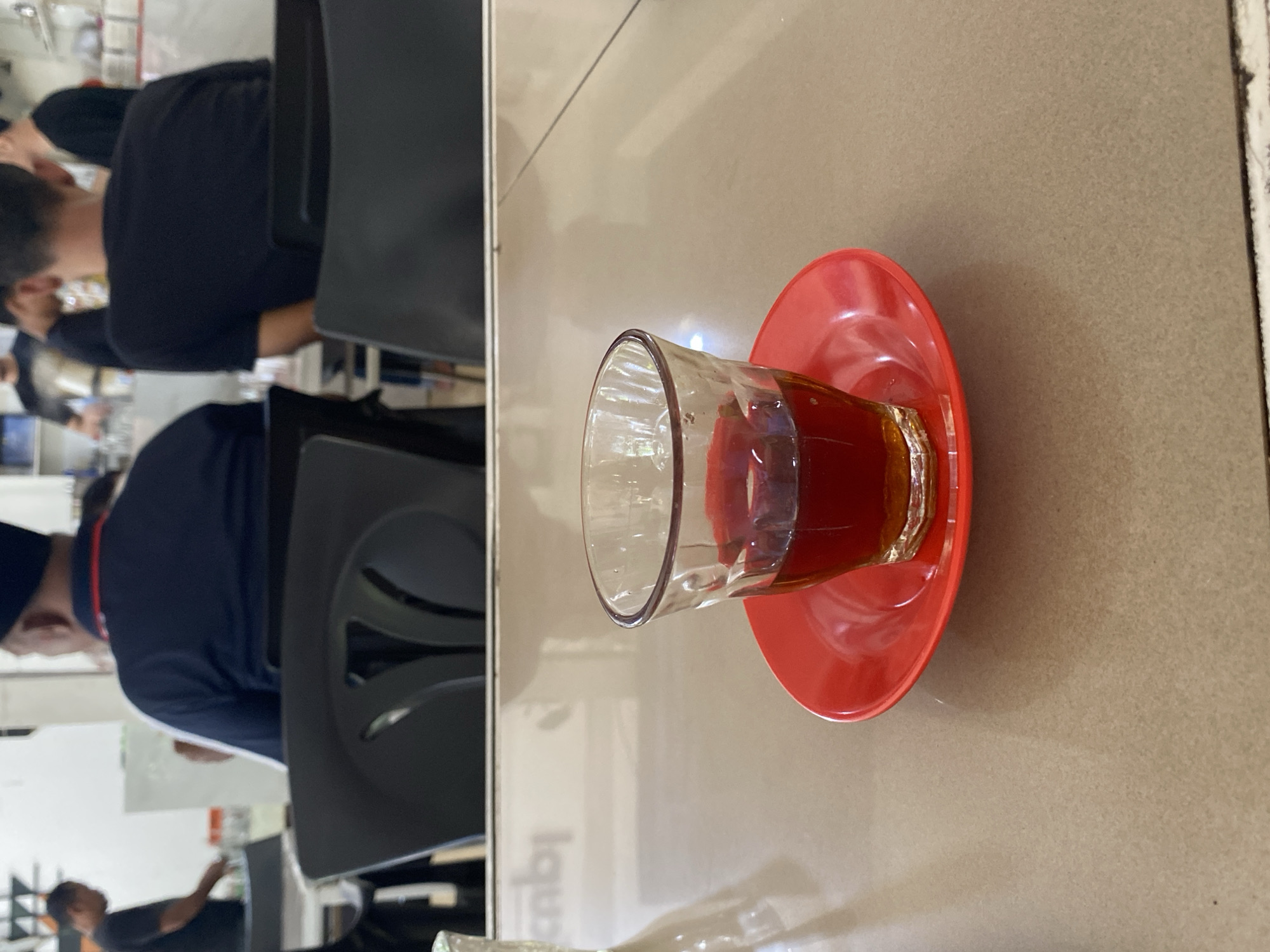 The image depicts a glass of tea on a red saucer, placed on a light brown table. In the background, several people are visible, some seated at tables and others standing. The setting appears to be an indoor establishment, possibly a cafe or restaurant, with white walls and shelves in the background. One of the individuals in the background is wearing a dark blue shirt and a black cap, and another is wearing a black t-shirt. The atmosphere suggests a casual gathering or meal. The scene is likely located in Lhokseumawe, Indonesia, a city known for its culinary scene and places where people gather for refreshments.
