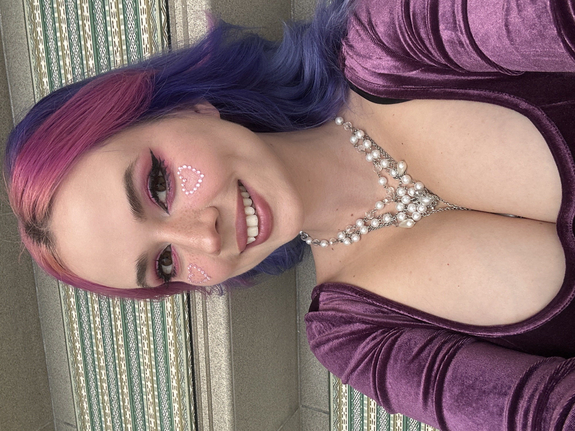 A close-up, angled portrait features a young woman with a joyful expression. Her two-toned hair transitions from bright pink at the front to vibrant purple. She wears elaborate eye makeup with pink eyeshadow and black eyeliner, complemented by small white heart-shaped rhinestones adorning her cheekbones, indicating a playful or festive mood. Freckles are visible across her nose and cheeks.
She is smiling broadly, revealing her teeth, and appears to be looking slightly off-camera. Her attire consists of a deep purple, long-sleeved velvet top or dress with a plunging neckline, suggesting a soft, luxurious texture. A prominent multi-strand necklace, made of pearls and silver-toned chains, drapes across her chest, adding an ornate touch to her ensemble.
The background is a subtly textured surface with vertical stripes in shades of green, white, and brown, possibly a patterned wall, tapestry, or a decorative panel. The lighting suggests daytime. No discernible activity beyond posing for the photograph is depicted. There is no visible text in the image. The setting is within the City of Zagreb, Croatia, although this information is external to the visual content of the image.