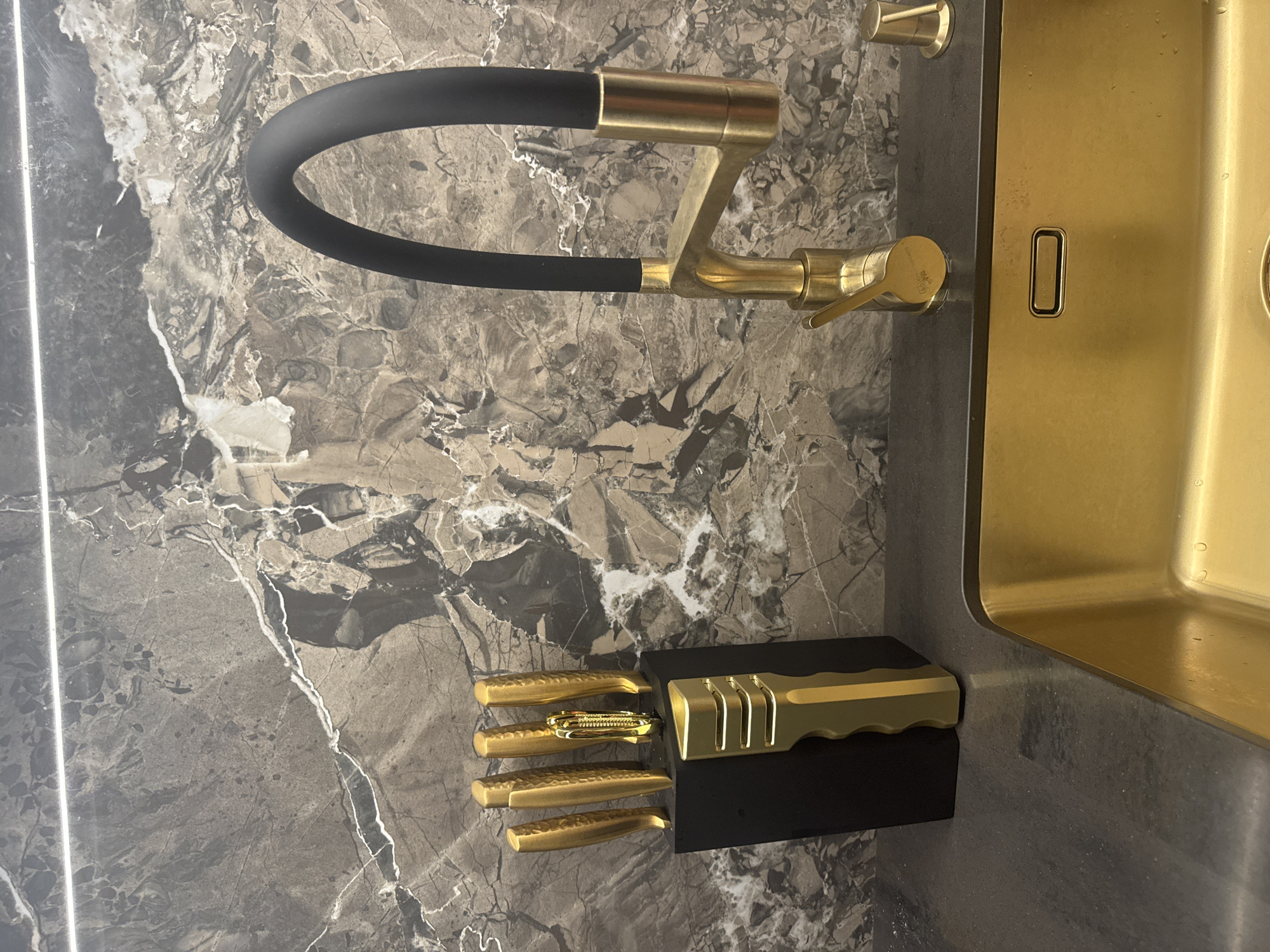 The image depicts a modern kitchen sink area with a prominent gold-colored sink and faucet. The faucet has a black hose and a gold-toned base and handle. Next to the sink, on a dark countertop, sits a black knife block holding several gold-handled knives. The backsplash is made of dark marble with white veining. The scene appears to be indoors, likely in a residential kitchen. There are no people visible in the image. The location is not specified to be in Sochi, Russia; this is an assumption.