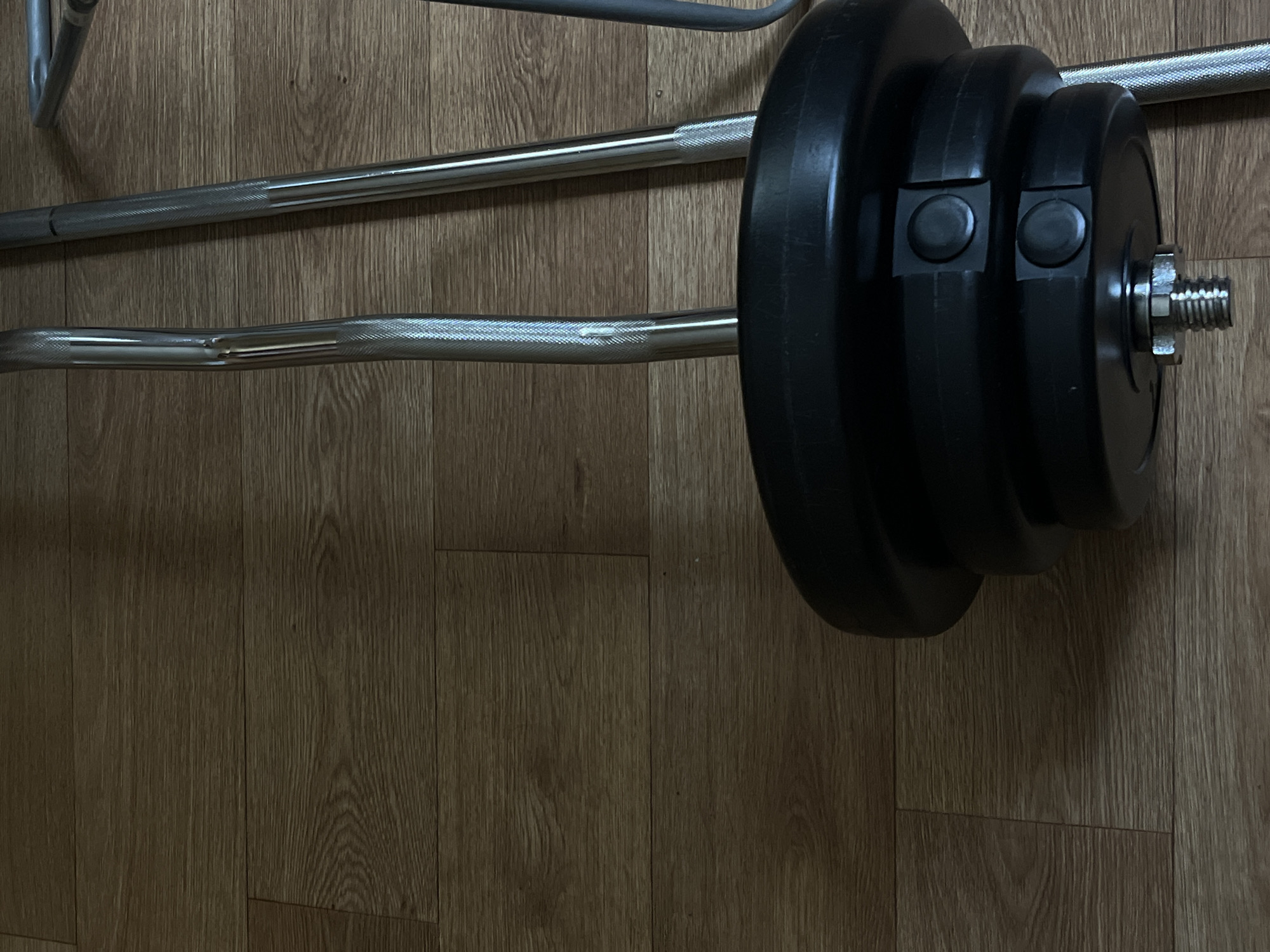 The image shows a close-up of weightlifting equipment on a wooden floor. In the foreground, a black weight plate with two circular black clamps is attached to the end of a barbell. The barbell, made of chrome-plated metal, has a knurled grip section. Behind this, another barbell, also chrome-plated, is visible. A third metallic bar, possibly part of a weight rack or another piece of equipment, is partially visible on the right edge of the frame. The wooden floor displays a wood grain pattern. There are no people present in the image. The scene depicts indoor exercise equipment in a domestic or gym setting. The specific location in Lviv, Ukraine, cannot be determined from the visual information provided.