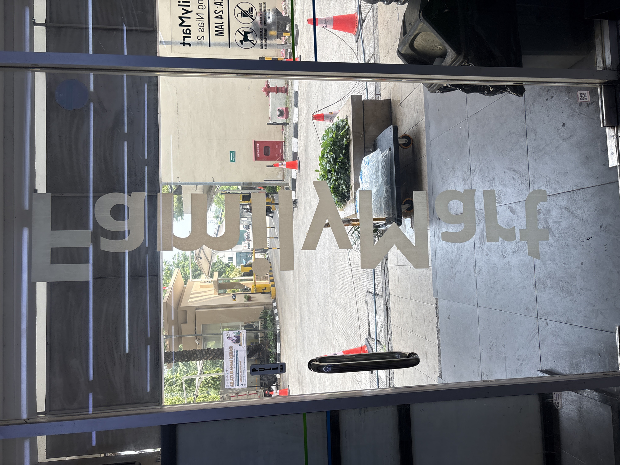 The image captures a view through a glass door, likely at the entrance of a commercial establishment. Prominently displayed on the glass are large, white, stylized letters that spell "TRUE MART" vertically. The letters are partially obscured by reflections from the environment outside.
The exterior scene visible through the glass shows a paved area, possibly a sidewalk or walkway, with some vegetation in planters. Orange traffic cones are placed strategically, one near a planter and another further in the background. A red fire hydrant is visible on the right side of the exterior, and a red utility box is mounted on a beige wall. Beyond this, a street scene with vehicles and buildings is implied, though not clearly detailed.
On the left side of the interior, near the glass door, a black door handle is visible. The floor inside appears to be tiled. On the far right, a black trash bin is present.
The text "MAL AS: A2" and "MALAYSIA" is visible on a sign on the exterior, suggesting the location might be related to Malaysia or have a Malaysian theme, which could be in North Jakarta, Indonesia, given the context provided. The overall impression is that of a retail space entrance, possibly a convenience store or a similar outlet.
