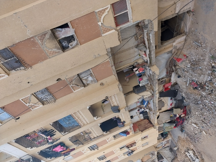 The image displays a multi-story residential building in Khan Yunis, Palestinian Territory, presenting extensive damage. The building's facade, predominantly beige with sections of faded orange, exhibits numerous cracks, impact points, and areas where plaster is missing. Many windows are shattered, with openings on various floors covered by black or white plastic sheeting, fabric, or blankets. Soot marks are visible around several window frames, particularly on the orange sections of the building. Laundry, including brightly colored clothes, hangs from multiple balconies, suggesting potential continued habitation or an attempt at normalcy. The lower levels of the structure are severely compromised, featuring fractured concrete slabs, exposed twisted rebar, and visible interior spaces. A significant amount of debris, including broken concrete, metal fragments, and various discarded items, litters the ground directly in front of and below the damaged sections of the building. Black plastic water tanks are observable on at least two lower-level balconies. The ground extending from the building is unpaved, covered in loose dirt and rubble. No individuals are discernible within the scene.