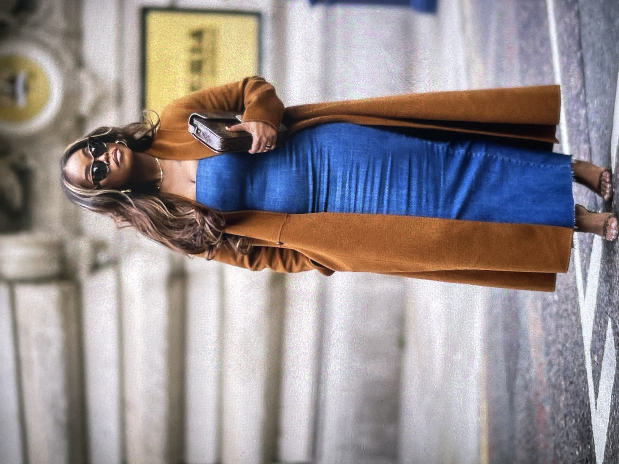 A full shot shows a woman with long, wavy brown hair and highlights, wearing sunglasses, a gold necklace, and gold hoop earrings, standing on a city street. She is wearing a strapless blue denim midi dress and a long, rust-colored coat. She is holding a dark brown clutch in her right hand and her left hand is in her coat pocket. She is wearing strappy, heeled sandals. The background shows a building with a yellow sign and a blurred street with white lines.