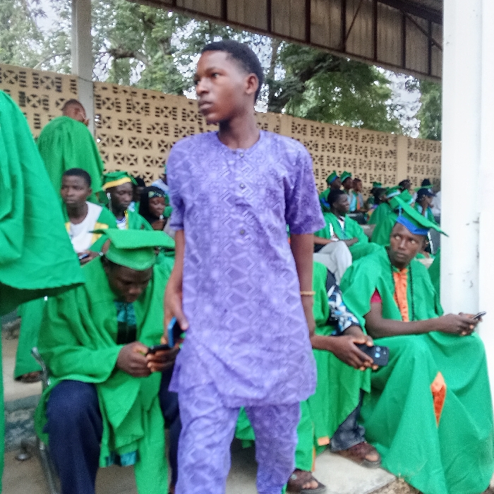 The image depicts an outdoor gathering of approximately twenty young adults under a concrete and metal-roofed shelter. In the foreground, a male, estimated late teens to early twenties, stands, facing right with his head slightly turned left. He wears a short-sleeved purple patterned tunic and matching trousers. A dark smartphone is partially visible in his left hand. A light-colored bracelet is on his right wrist. Around and behind him, multiple individuals, predominantly male, are seated on tiered concrete benches, all wearing green academic gowns and mortarboard caps, some with blue or yellow accents. Several seated individuals are engaged with black smartphones; one in the lower-left is looking intently at his phone, while another to the right of the standing figure holds a phone and looks forward. The background features a light-colored wall with geometric open patterns, beyond which trees and foliage are visible under a light sky. White pillars supporting the shelter show signs of wear. The exact location within the city of Akwanga, Nigeria, cannot be determined from the visual information provided.