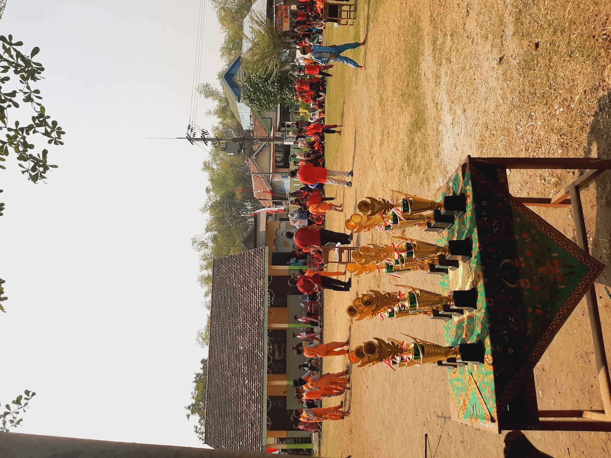 The image captures an outdoor event, likely a school sports day, taking place in the village of Sumberjo, Indonesia. In the foreground, a table is adorned with a green, patterned cloth, upon which several golden trophies are displayed. Behind the table, a crowd of people, primarily children dressed in orange outfits and adults in red and white attire, are gathered on a dry, grassy field. The backdrop features a long, single-story building with a tiled roof, characteristic of a local school or community hall, and several other houses. The overall atmosphere suggests a sunny day with a clear sky, a common occurrence in the region.