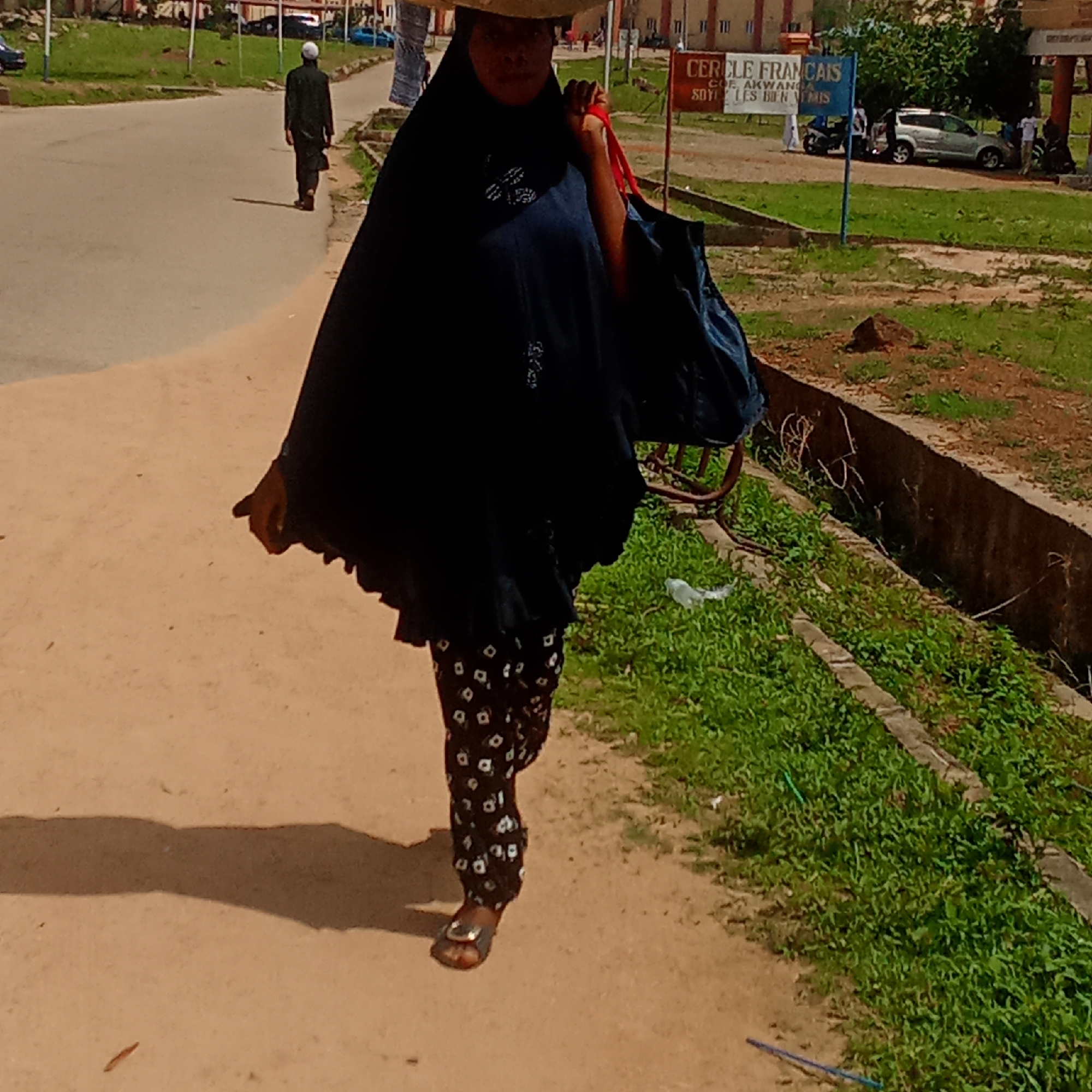 The image captures a sunlit street scene in Akwanga, Nigeria, featuring several individuals and elements of an urban environment. In the foreground, a woman walks along a sandy path bordering a paved road. She wears a dark blue or black flowing garment with white embroidered patterns, patterned trousers, and sandals. She carries a red-strapped bag over her shoulder and appears to have a wide-brimmed hat on her head, partially obscuring her face.
Further down the paved road, a man dressed in dark attire and a white skullcap walks away from the camera, casting a noticeable shadow, suggesting a bright, sunny day. The road appears to curve slightly. To the right, there's a strip of green grass and a concrete drainage channel.
In the mid-ground on the right side, a blue and white sign is prominently displayed, bearing the French text: "CERCLE FRANCAIS COE. AKWANGA SOYI LES BIEN VENUES". Beyond the sign, a silver car, a motorcycle, and several other people are visible near some buildings, indicating activity. The overall setting is an open-air public space, likely a street or pathway within a community.