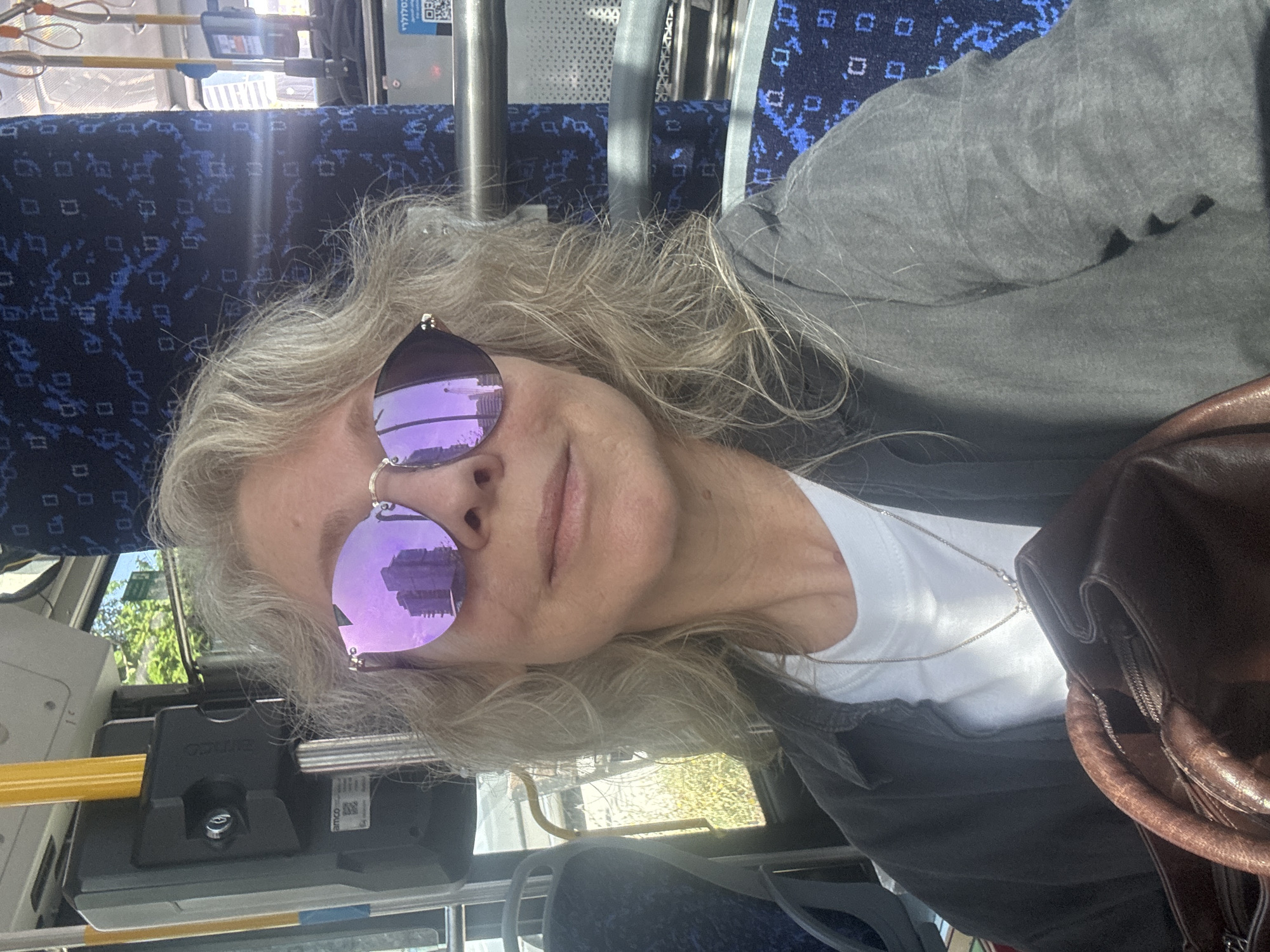 A person is sitting on a bus. The bus has blue patterned seats and metal handrails. The person is wearing sunglasses with pink lenses, a white shirt, and a gray jacket. A brown bag is next to the person. The reflection in the sunglasses suggests a cityscape, possibly in Tel-Aviv, Israel. There is a device on the left with a yellow handle, and possibly an electronic display above it. The lighting is bright, suggesting it's daytime.