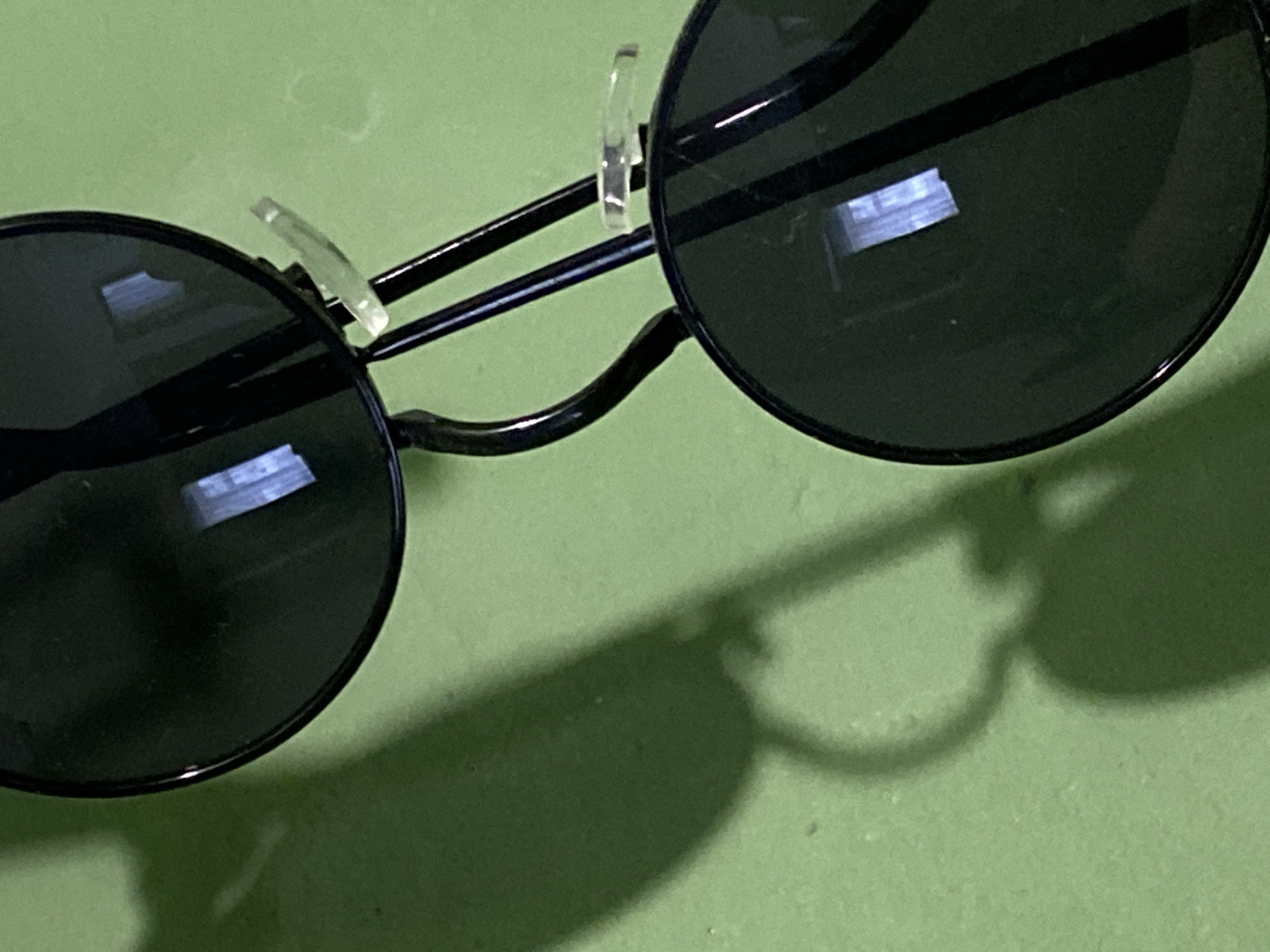The image displays a close-up, oblique view of a pair of black-framed sunglasses with dark, circular lenses, positioned against a textured, light green surface. The sunglasses are partially visible, angled diagonally from the bottom-left to the upper-right of the frame. Two clear, rectangular reflections of a light source or window are present on each of the visible lenses. Transparent nose pads are visible on the bridge of the sunglasses. A distinct, dark shadow of the sunglasses, including the frame and temple arms, is cast onto the green background, extending from the bottom-left corner and running parallel to the physical object. The green surface appears to be a wall or flat panel. No individuals or direct actions are depicted. The lighting is strong, creating defined shadows and reflections. The precise location within the city of Chaikovsky, Russia, cannot be determined from the visual information provided.