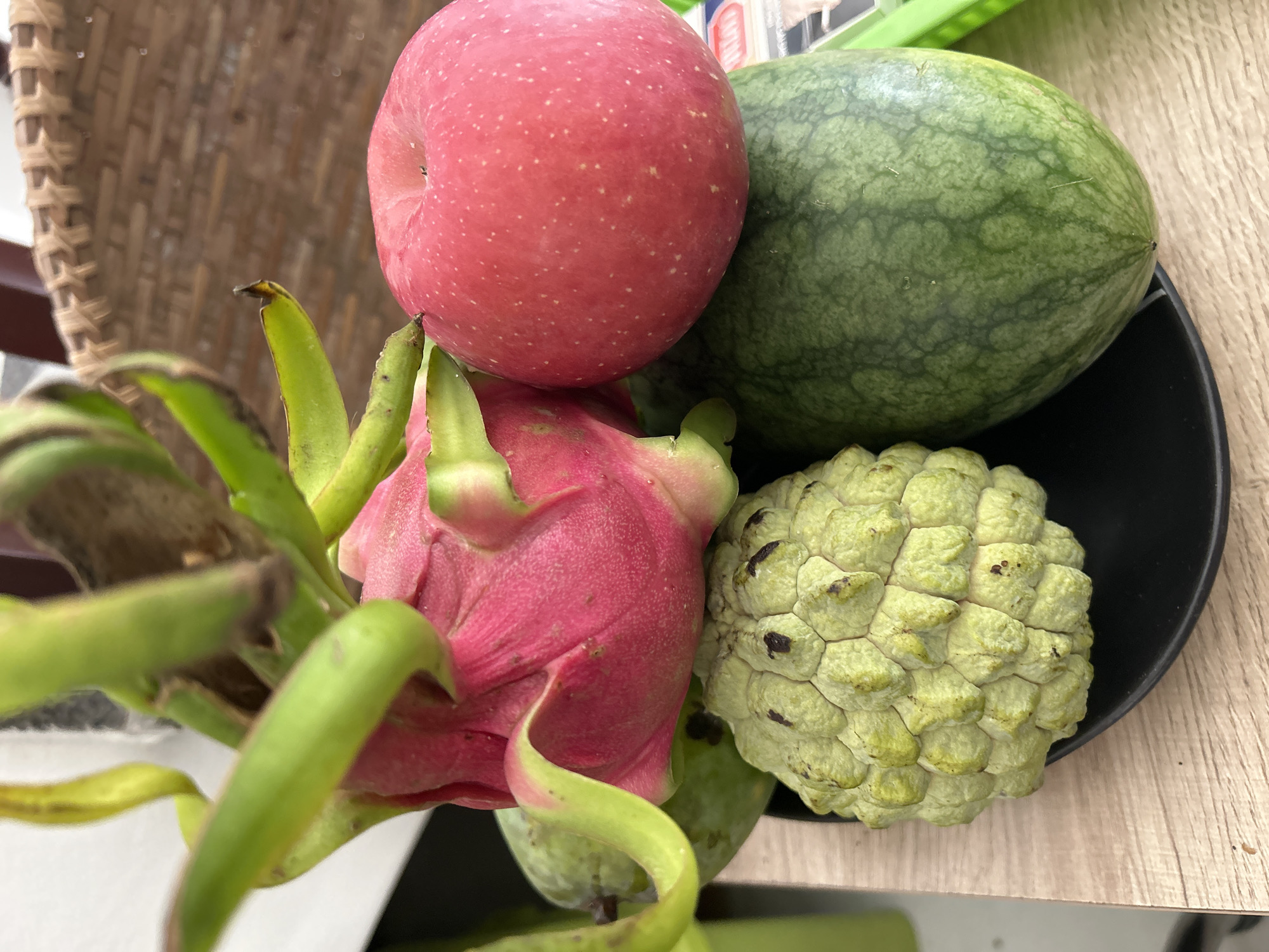In Nha Trang Ward, Vietnam, a close-up shot captures a still life arrangement of assorted fruits on a black plate, resting on a light-colored wooden surface. The collection includes a vibrant pink dragon fruit with green, leafy bracts, a red apple with faint white speckles, a textured, light green soursop, and a dark green watermelon with lighter green stripes. The fruits are stacked and positioned to create a visually appealing display. The background is blurred but suggests an indoor setting with a woven basket visible.