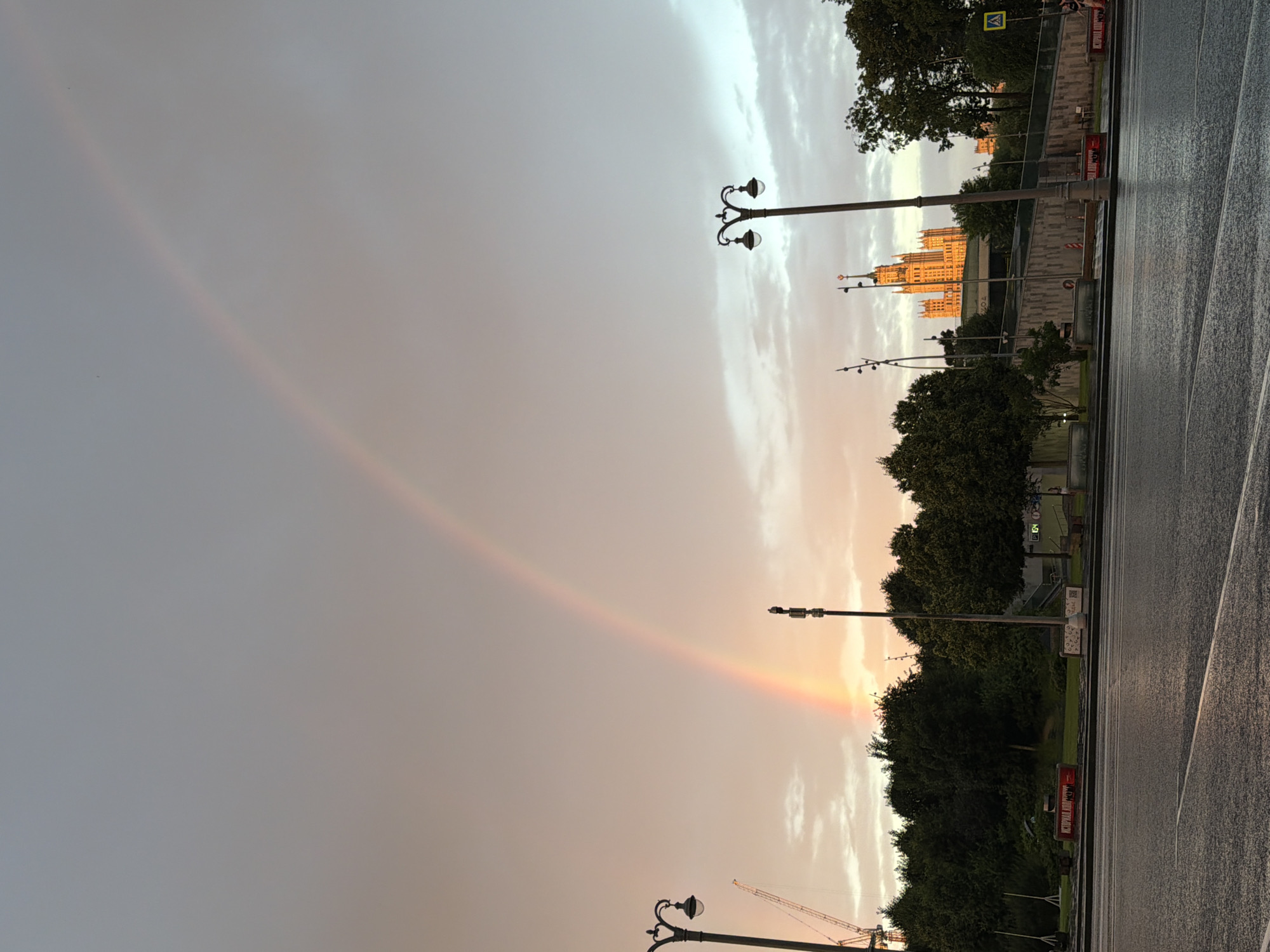 The image depicts a cityscape scene in Moscow, Russia, at dusk, as evidenced by the soft, diffused light and the presence of a rainbow arching across the sky. The foreground features a wet asphalt road, indicating recent rain. To the left, a lamppost stands next to a construction crane, suggesting ongoing development or renovation. A dense line of trees forms the middle ground, with a distinctive Stalinist skyscraper, illuminated by the warm sunset light, dominating the background on the right. Several other lampposts and streetlights are visible throughout the scene, contributing to the urban ambiance. The sky is overcast with patches of lighter clouds, and the rainbow is a prominent feature, adding a touch of natural beauty to the urban landscape.