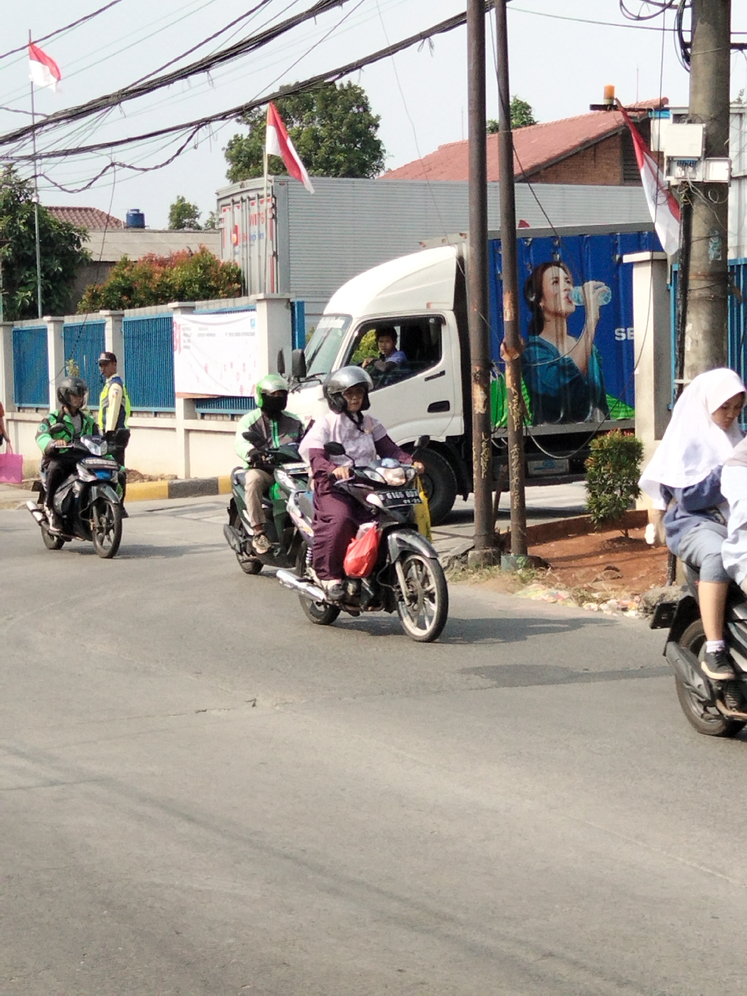 This image captures a street scene in South Tangerang, Indonesia, likely during the daytime. Several individuals are riding motorcycles on a paved road. In the foreground, a person wearing a white hijab and a dark shirt is riding a motorcycle. Further ahead, two individuals are riding motorcycles, one wearing a green helmet and jacket, and the other a silver helmet and a pinkish-purple top. To the left, another person on a motorcycle is visible. Behind the motorcyclists, a white truck with a blue container is parked. The container features a large advertisement depicting a woman drinking water. Red and white Indonesian flags are visible on poles in the background, along with electrical wires and buildings. The environment appears to be urban or semi-urban, with some trees and foliage present.