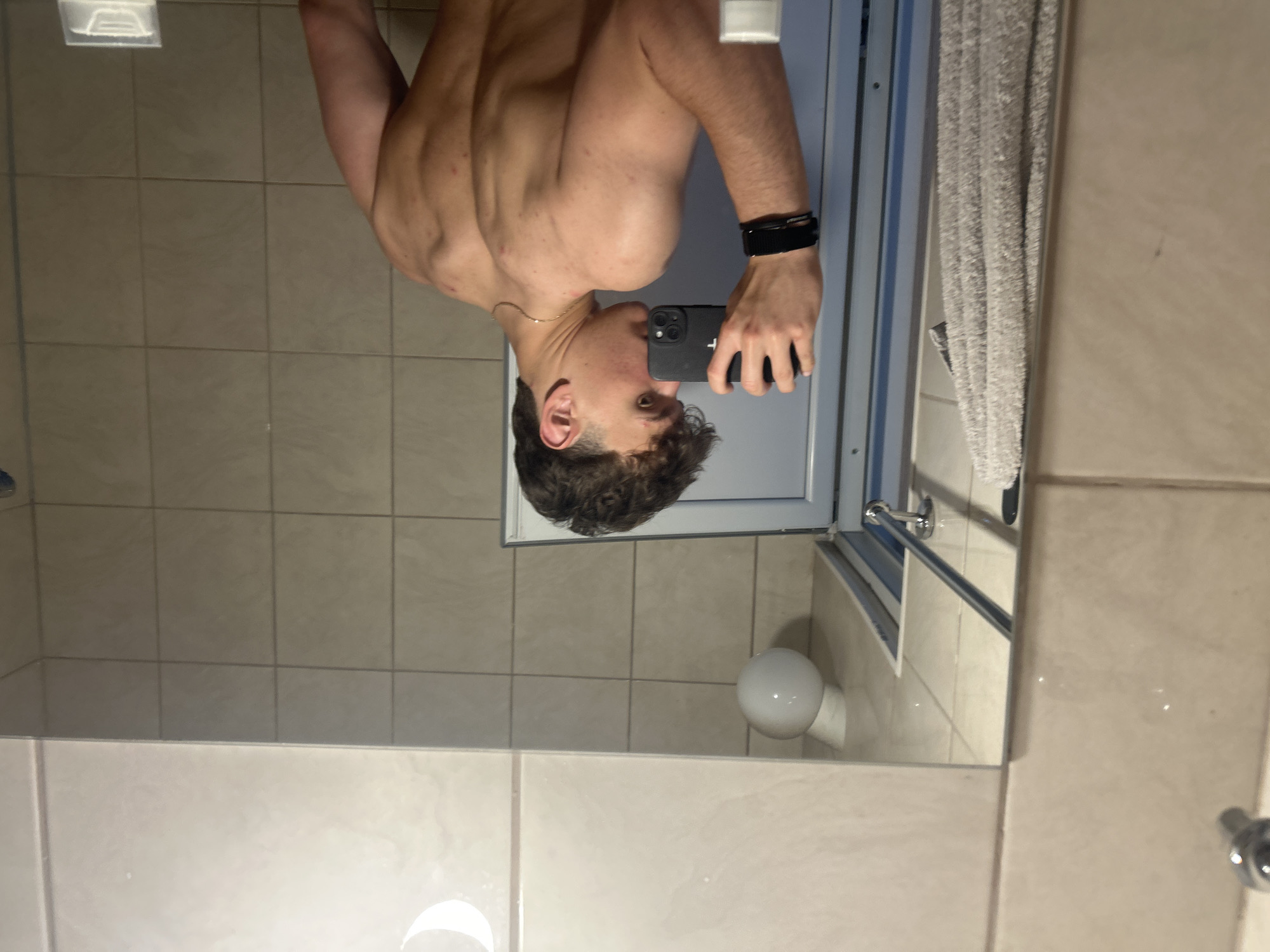 A photograph shows a shirtless person holding a smartphone in a bathroom. The person's back and shoulder muscles are visible. They are wearing a black wristband on their left wrist and a gold chain around their neck. The smartphone appears to be used to take a picture in a mirror. The bathroom has tiled walls, a towel hanging on a rack, a shower door with a chrome handle, and a light fixture. The specific location is Unknown, Unknown.