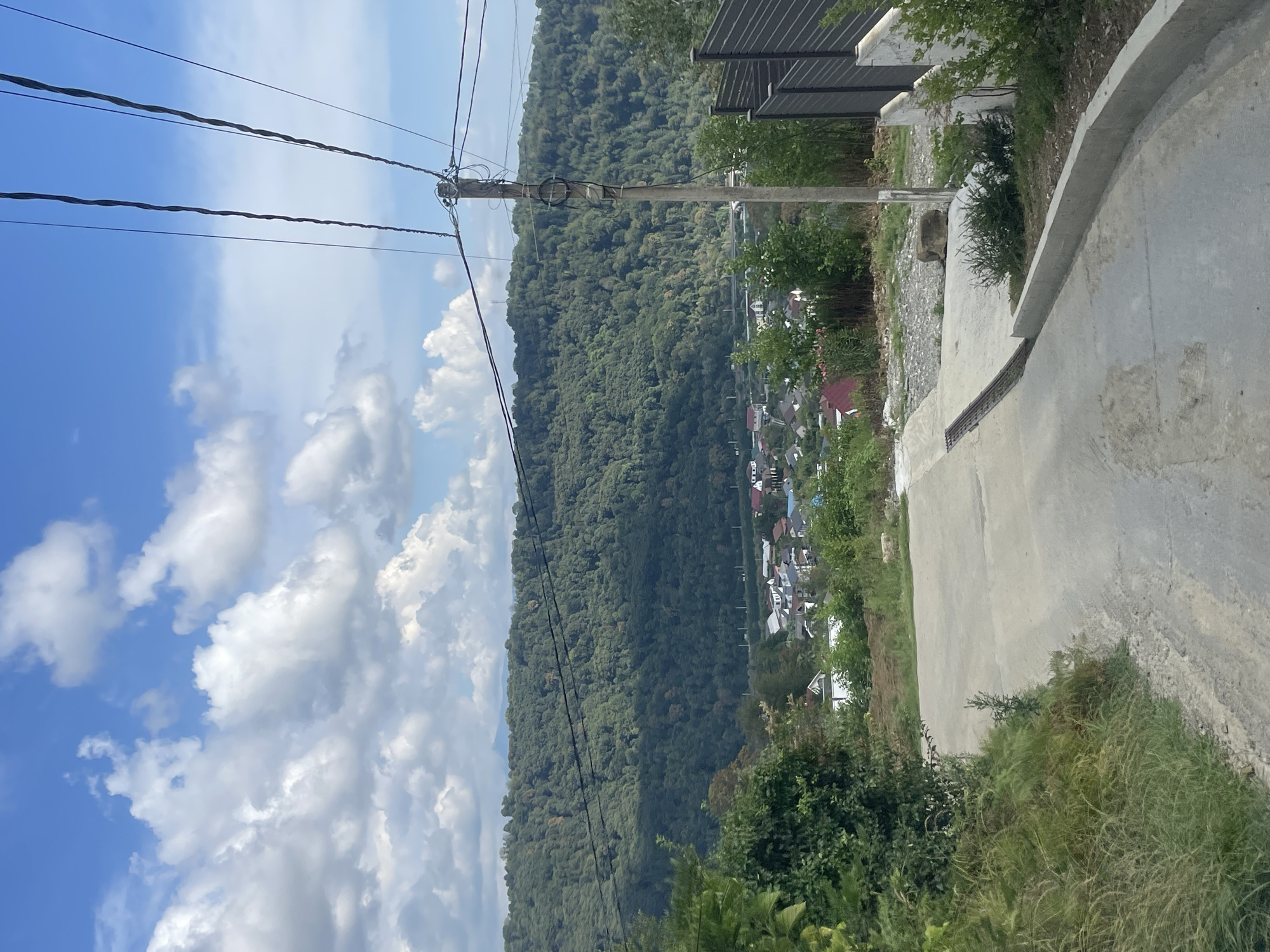 This image depicts a descending paved road in a rural or suburban area. A wooden utility pole with multiple wires runs vertically through the right side of the frame. In the background, a densely forested hill rises, with a scattering of houses visible at its base. The sky is blue with scattered white clouds. The area appears to be the outskirts of the city of Vysokoye, Russia, a location known for its mountainous terrain and lush vegetation. There are no people or animals visible in the image.