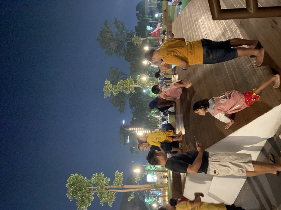 A nighttime scene is depicted in an outdoor public space in Sidoarjo, Indonesia, illuminated by artificial lighting from lampposts and decorative vertical light strips. The environment features multiple trees with green foliage and sloped wooden recreational structures. In the immediate foreground, a man in a black t-shirt and light-colored shorts, wearing flip-flops, stands on a flat white concrete surface, engaged with a smartphone. Adjacent to him, a young girl with dark hair, wearing a pink and white jacket over red patterned shorts, barefoot, is positioned on a sloped wooden surface, leaning forward. To the right, another man in a yellow t-shirt and dark blue shorts, barefoot, stands facing away from the camera on a sloped wooden ramp, holding a smartphone with both hands. In the mid-ground, a woman crouches on a flat surface, smiling towards the camera, alongside a small child in a yellow patterned outfit who is also looking towards the camera. Various other individuals, including adults and children, are visible throughout the scene, some sitting or playing on the wooden structures and grass areas. A red tent structure is discernible in the distant background on the right. The sky is dark blue, indicating night.