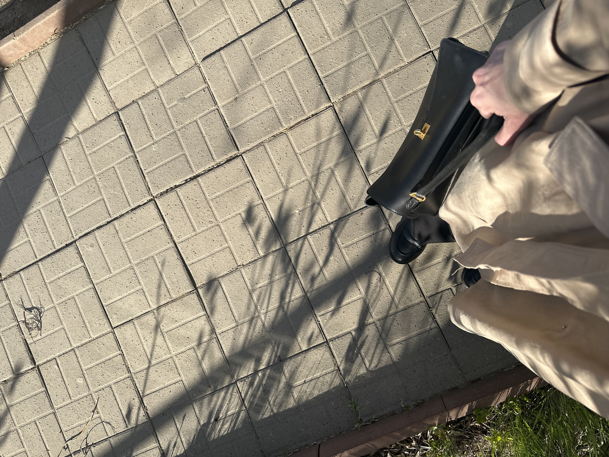 The image depicts a section of a paved walkway in Chaikovsky, Russia. The walkway is constructed from light-colored, rectangular paving stones arranged in a basketweave pattern. Long shadows fall across the walkway, suggesting a low sun angle. A person, only partially visible, is wearing a light-colored trench coat and dark shoes. They are carrying a black leather handbag, which they appear to be opening. The scene is outdoors and appears to be taken during daylight hours. There is grass and vegetation visible near the edge of the walkway.