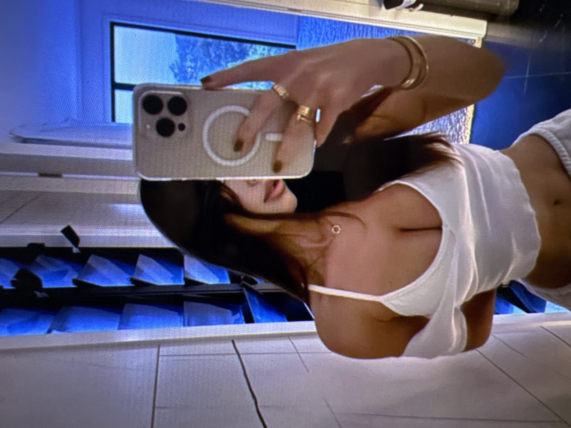A woman is taking a mirror selfie in a room with blue lighting. She is wearing a white top and has long dark hair. She is holding up a cell phone in her right hand, and she is wearing rings on her fingers and bracelets on her arm. The room appears to be a bathroom, with tiled walls and a door in the background. The lighting creates a surreal and abstract atmosphere. There is no information to suggest this location is in Castelo Branco, Portugal.