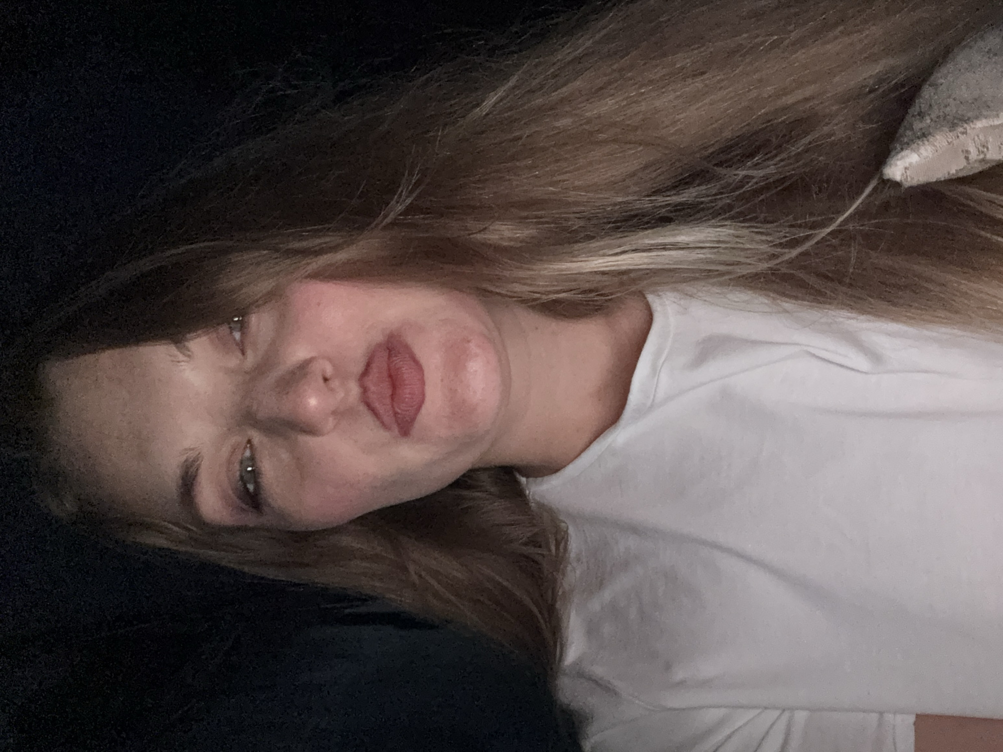 A close-up, frontal, waist-up selfie of a woman with long, blonde hair and fair skin is taken in an indoor setting with dark surroundings. The woman is wearing a white t-shirt and is looking directly at the camera with her lips pursed. Her eyes are partially closed, and she has light makeup on her face. A patterned pillow is visible on her right side, partially obscured by her hair. There is no discernible information about the location in Denpasar, Indonesia.