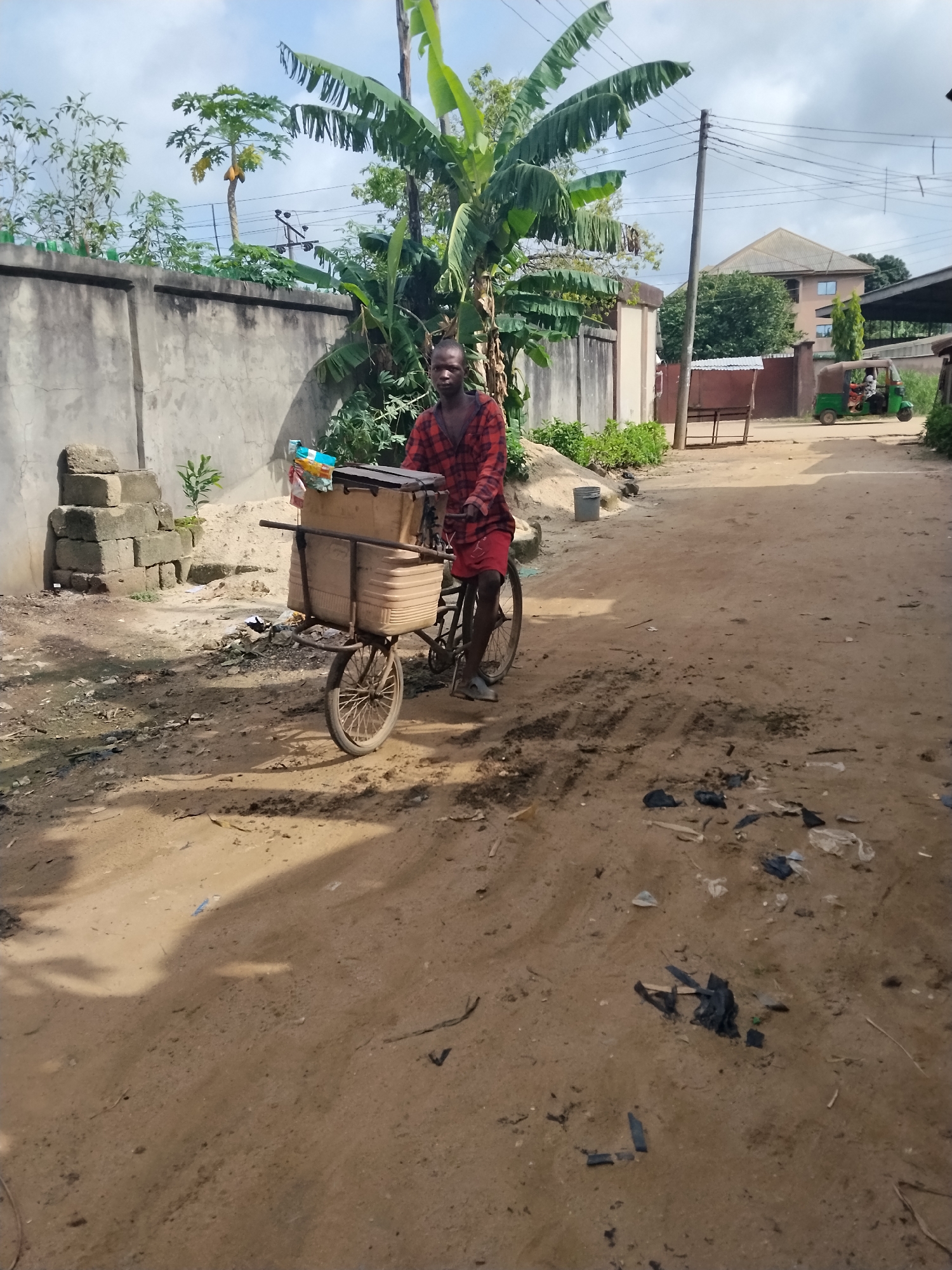 The image depicts a man riding a bicycle cart on a dirt road in what appears to be a residential area. The man is wearing a red plaid shirt and shorts. The cart is loaded with boxes. The environment is characterized by a concrete wall, tropical vegetation, and scattered debris on the ground. In the background, a building and a three-wheeled vehicle (tuk-tuk) are visible. The location is inferred to be in Umuode, Nigeria.