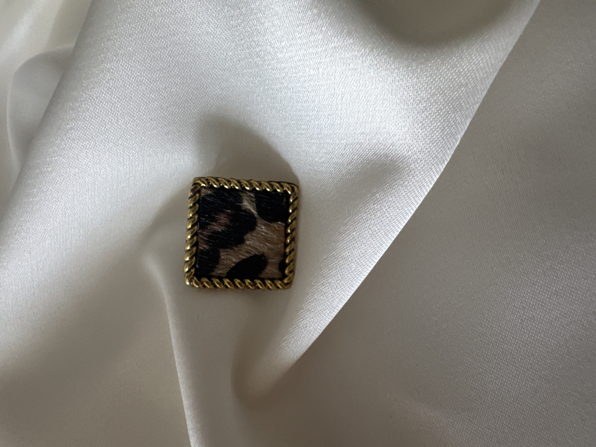 The image shows a square-shaped ring with a gold twisted rope border. The center of the ring features a leopard print pattern with brown, black, and tan colors, resembling fur. The ring is placed on a white satin fabric, which is draped and creased, creating soft highlights and shadows. The overall scene is a close-up studio shot. The image does not contain any information about the city of Bishkek, Kyrgyzstan, or any people, actions, or interactions.
