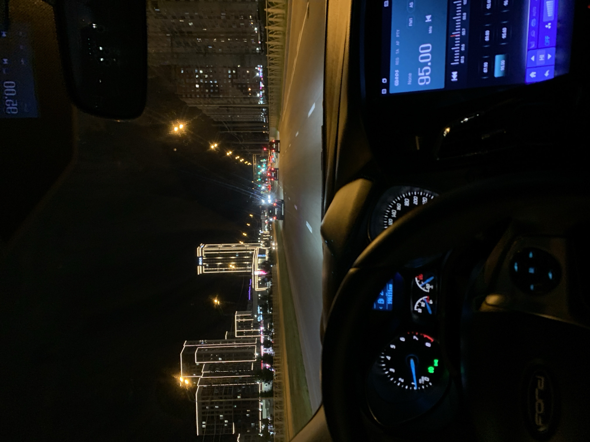 The image depicts a night-time view from the interior of a moving car on a multi-lane highway in Stavropol, Russia. The foreground shows the steering wheel and dashboard of the vehicle, including a digital display showing "95.00" for radio frequency and a speedometer. The road ahead is illuminated by streetlights and headlights of other vehicles. Tall buildings with modern architecture, lit up with decorative lights, are visible on the left side of the road, characteristic of the newly developed residential areas of Stavropol, such as the area near the "Nevinnomyssky Azot" plant or the "Symphony" residential complex. On the right side, apartment buildings with illuminated windows are also visible. The sky is dark, indicating it is nighttime. The overall scene suggests urban driving through a well-lit part of the city at night.