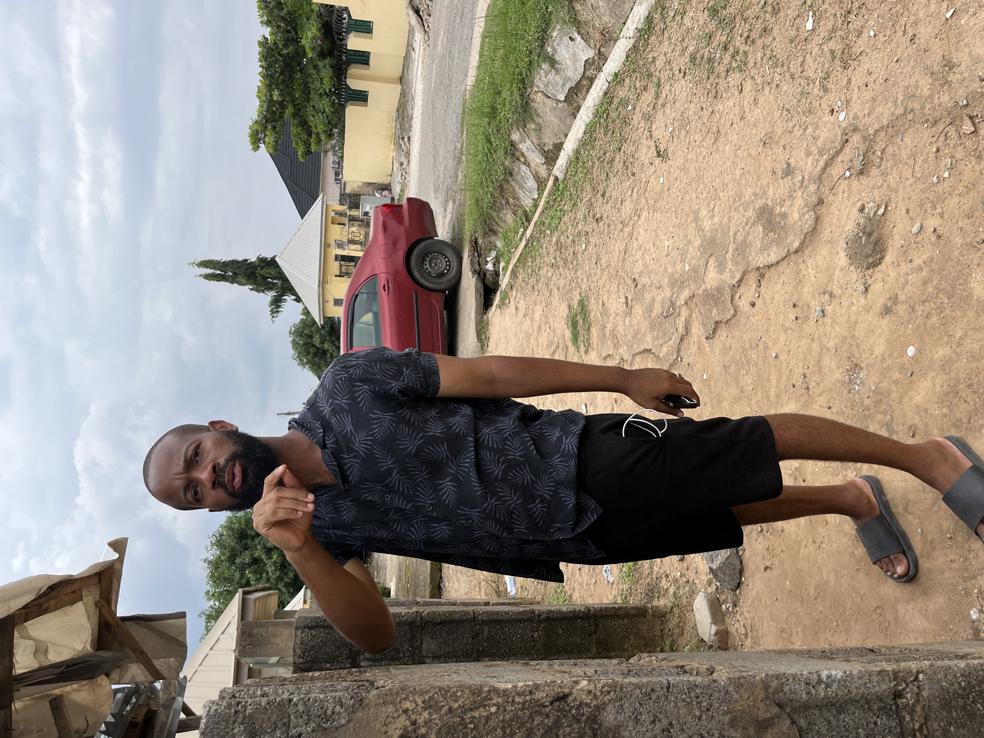A full-body, medium shot captures a Black man with a beard standing outdoors, facing forward and slightly to the left, in what appears to be a residential area in Abuja, Nigeria. He is wearing a short-sleeved, black collared shirt with a gray leaf pattern and black shorts. His left hand is raised to his chin, and his right hand is by his side, holding a black object. He is wearing black slide sandals. The man stands on a dirt path next to a rough, concrete structure. In the background, a red sedan is parked on a paved road. Beyond the road, there are yellow and light-colored buildings, some with pitched roofs, and lush green trees. The sky is overcast with gray clouds. A narrow, vegetated drainage ditch runs along the side of the road. The overall impression is a casual, everyday scene in an urban environment with modest infrastructure.