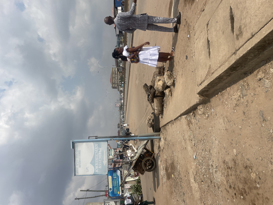 The image displays an urban street scene in Kwashieman, Accra, Ghana, identifiable by a prominent sign reading "CHRIST APOSTOLIC CHURCH INTERNATIONAL KWASHIEMAN ASSEMBLY." The foreground features a rough, unpaved dirt shoulder with scattered debris, transitioning into a damaged concrete curb and an exposed drainage channel.
To the left, a blue-framed sign stands erect, positioned next to a discarded, rusted metal pushcart or wheelbarrow that appears partially loaded. Immediately to the right of the sign, a pile of large rocks, several cut logs (possibly palm tree trunks), and a single black tire are stacked on the dirt ground.
On the right side of the frame, two individuals stand on the dirt shoulder. A woman, dressed in a white sleeveless dress and flat sandals, carries a small child secured on her back with a patterned fabric wrap. She is observed looking towards the paved road. Adjacent to her, a bald man wearing a patterned grey and white suit and dark shoes stands with his back mostly to the camera, extending his right arm and hand in a gesturing motion towards the street.
In the mid-ground, a multi-lane paved road extends into the distance, with several vehicles and pedestrians visible. Streetlights line the road, and a speed limit sign indicating "50" is discernible. The background features various commercial or residential buildings, including a multi-story structure on the far side of the road. The sky above is heavily overcast with dense grey clouds. A person in a green outfit is partially visible on the far left, near the pushcart.