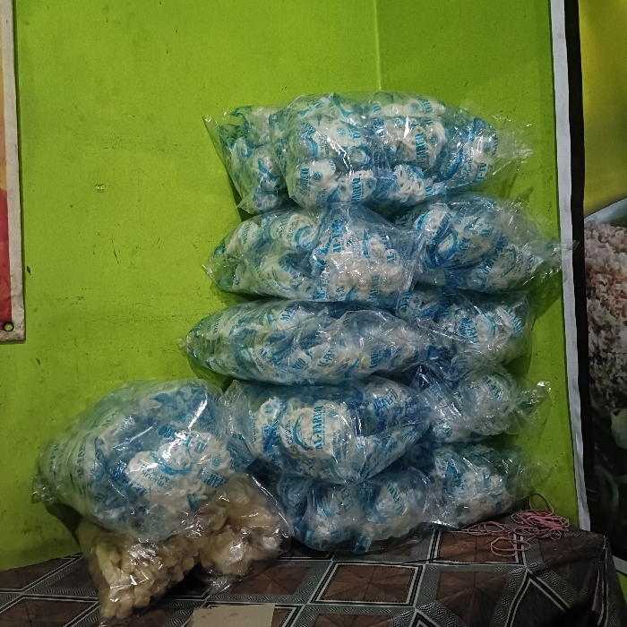 The image shows a stack of clear plastic bags filled with white, round objects, likely candies or sweets. There are at least 8 bags in the stack, all showing the same packaging with blue lettering. The scene is set against a green wall. The bags are on a table, partially covered by a brown and white patterned cloth. The table is also covered in some items of white fabric and a few rubber bands. The image was taken at coordinates -6.975 latitude and 110.332 longitude, which is in the city of Tugu, Indonesia.
