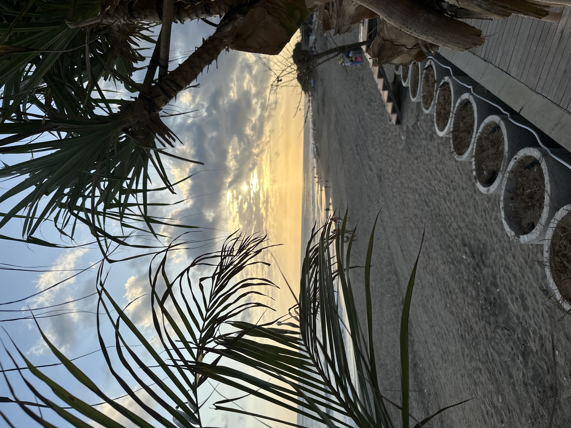 The image captures a sunset scene at a beach in North Kuta, Indonesia. A sandy beach stretches into the distance, bordered by the ocean and a pedestrian walkway lined with decorative concrete planters. Palm trees are positioned on the left side, framing the view of the sunset. The sun is partially obscured by clouds, casting a golden light across the sky and reflecting on the water. Some people can be seen as small figures on the beach.