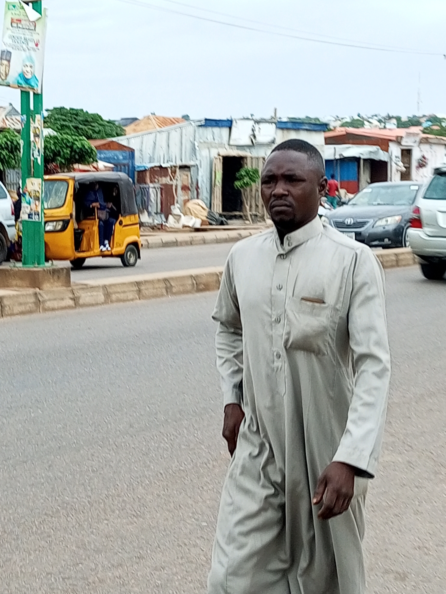 The image shows a man walking on a paved road, in what appears to be a market area in Tempure, Nigeria. He is wearing a light-colored, loose-fitting garment with a high collar and buttons down the front. The man has dark skin and short, dark hair, and his expression is neutral, looking slightly off-camera.
In the background, there are several vehicles, including a yellow auto-rickshaw with a driver and passenger. Behind the auto-rickshaw, there are various market stalls and buildings made of corrugated metal and other makeshift materials, suggesting a busy commercial area. A few trees are visible, and the sky is overcast, indicating a cloudy day. There is a pole on the left with some posters or advertisements attached to it. One poster shows portraits of two individuals. The overall scene depicts a typical day in a bustling Nigerian town.