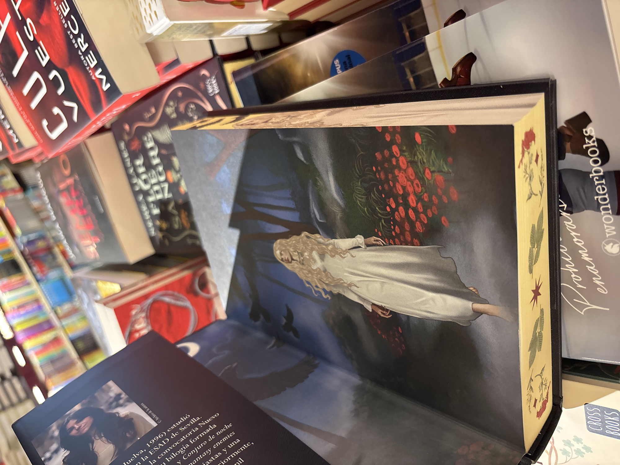 The image is a close-up, eye-level shot of several books displayed in a store, likely a bookstore. In the foreground, a book is open, revealing an illustration of a woman with long blonde hair wearing a flowing white dress, standing in a field of red flowers. The background shows shelves filled with more books. The overall lighting suggests an indoor setting, possibly a market or a shop. One of the visible book titles is "CULPA VUESTRA" by MERCE. Another is "FLOR DE SANGRE". The scene does not provide specific details that would allow for its localization in the city of Orihuela, Spain, or any other specific city.