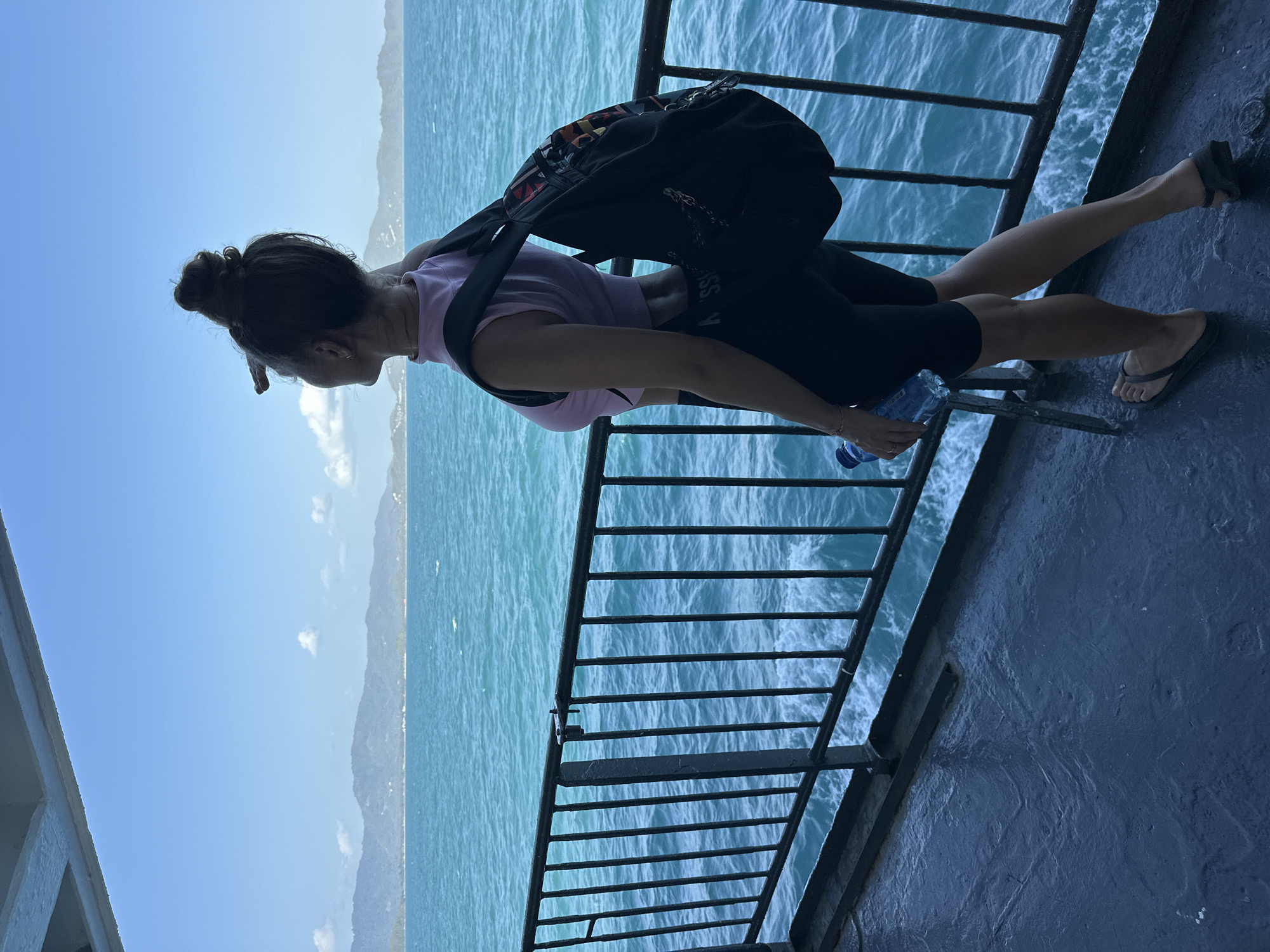 A woman stands on the deck of a ferry, looking out at the ocean. She is wearing a light pink crop top, black shorts, and flip-flops. She has a black backpack on her shoulders and is holding a bottle of water. The ferry is moving, and the water is a vibrant blue. In the distance, there are green hills and a clear blue sky. The deck of the ferry is wet and dark, and there is a black metal railing to her left. The scene appears to be in Thailand, possibly on a trip to one of its many islands.