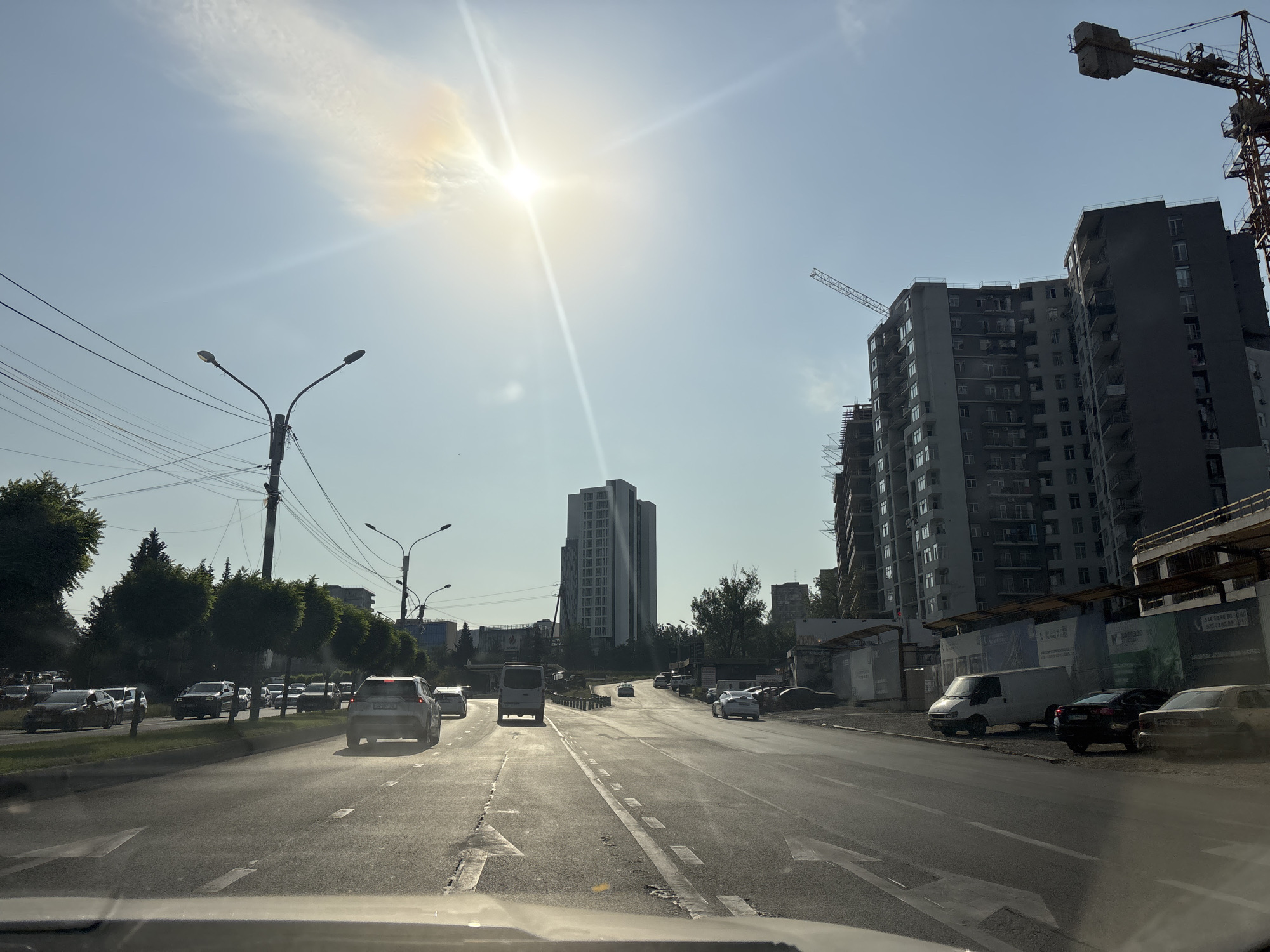 The image depicts a sunlit street scene in Tbilisi, Georgia, featuring a multi-lane road with moderate traffic. The perspective is from a vehicle, looking forward. On the right side of the road, there are several multi-story buildings under construction, characterized by scaffolding and cranes. One prominent building is dark gray, with many windows. To the left, a row of green trees lines the road, with a tall, modern white building visible further back. Overhead, a bright sun with visible rays is present in a clear blue sky, with a faint halo effect around it. Streetlights and power lines are also visible. The road itself has clear lane markings, and several cars, including a white van and a silver SUV, are in motion or parked. The overall impression is an urban environment experiencing ongoing development under a bright, sunny sky.
