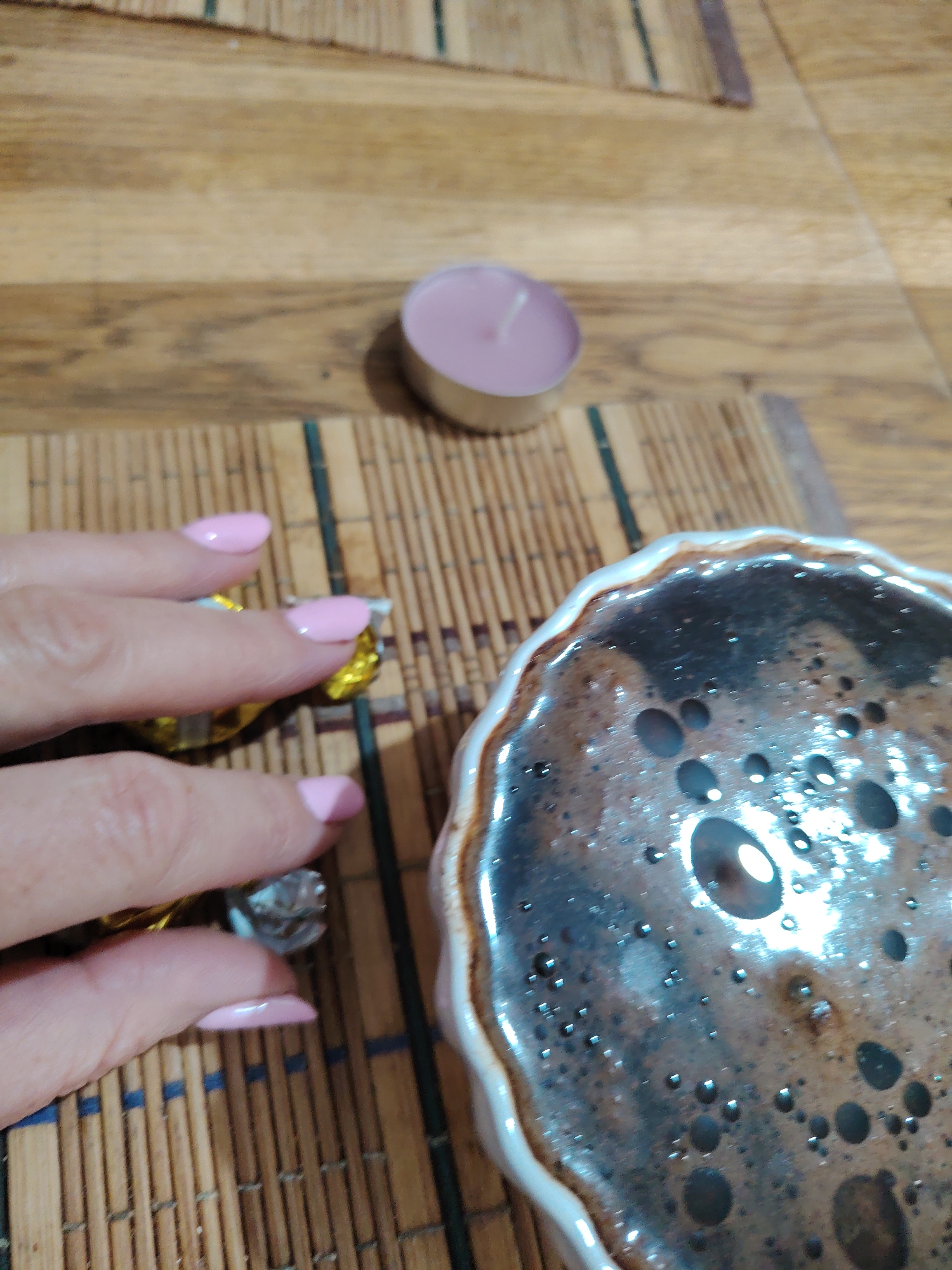 A close-up, over-the-shoulder shot shows a person's hands with light pink painted fingernails holding two individually wrapped candies. The candies are wrapped in gold foil. To the right of the hands, a ceramic dish containing a dark liquid with a frothy surface is visible. The dish has a wavy, cream-colored rim and a speckled brown and black interior. In the background, a small, unlit pink tea light candle sits on a wooden surface. The wooden surface appears to be a placemat made of thin bamboo strips. The scene is indoors, on a table. No specific location within Hrodna, Belarus can be determined from the visual information provided.
