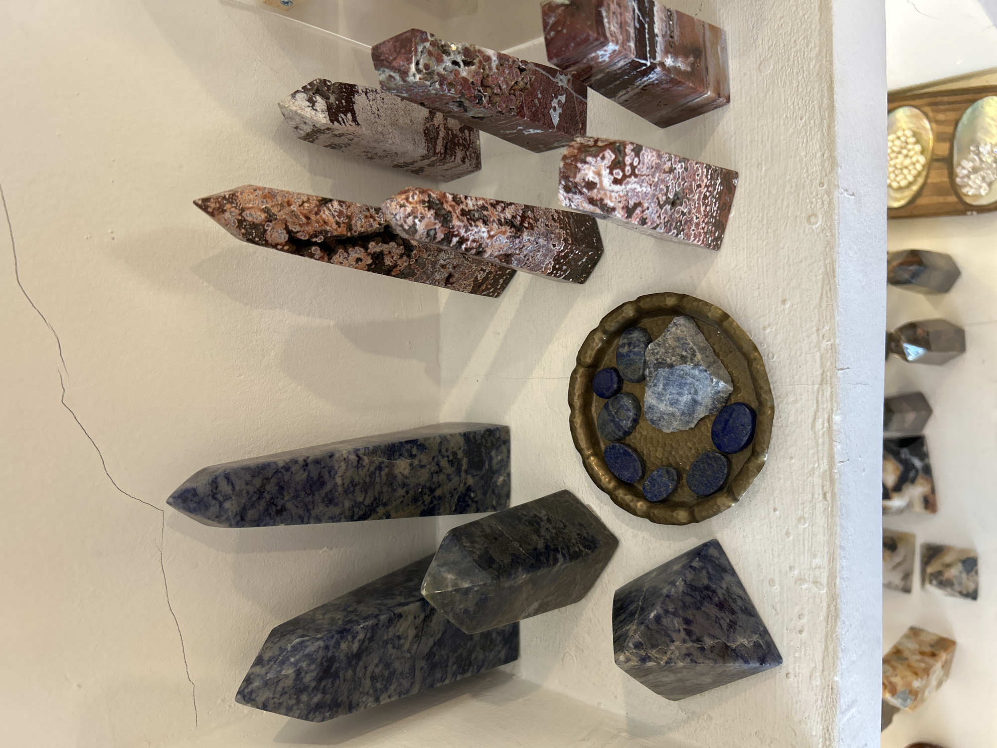 The image shows a collection of polished gemstones displayed on a white shelf. The gemstones are primarily in the shape of obelisks and pyramids, with some smaller tumbled stones. There are two main types of gemstones visible: a dark blue stone with white veining, and a reddish-brown stone with white and black inclusions, resembling jasper or agate. The blue stones are arranged on the left side of the shelf, with a pyramidal stone in the foreground and several obelisks behind it. The reddish-brown stones are on the right side, also displayed as obelisks of varying sizes. In the center of the shelf, there is a small, ornate brass dish containing several small, dark blue, oval-shaped stones and a larger, rougher piece of a similar blue stone. The background is a white wall with a visible crack running across the top. The shelf itself is also white and appears to be made of wood. There are no people visible in the image. This arrangement of polished gemstones is typical of those found in tourist shops or gemstone galleries, commonly sold in areas like Sukawati, Indonesia, which is known for its art markets and artisanal crafts.