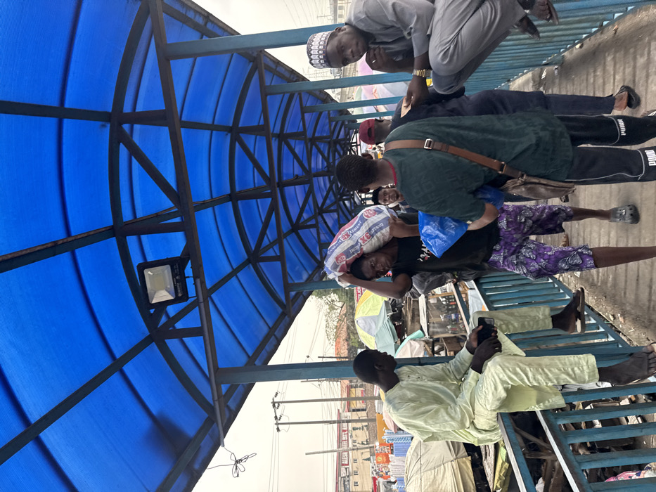 This image depicts a segment of a covered pedestrian walkway or bridge in Lekki, Nigeria, specifically above or adjacent to a bustling market area. The walkway features a blue translucent corrugated roof supported by a dark metal arched framework and blue metal railings. An overhead floodlight is mounted on the roof structure.
In the left foreground, a male individual wearing a light green traditional outfit (tunic and trousers) is seated on the blue railing, facing right, looking down at a smartphone held in his hands. His feet are bare.
Mid-ground, moving from right to left, a male individual with dark skin wears a black t-shirt and purple patterned shorts. He carries a large white and red striped sack, marked "KGI", over his right shoulder and a blue plastic bag in his left hand. He wears dark clogs. Directly behind him, another male individual is seen from the back, wearing a dark green t-shirt, black Puma track pants, and a brown crossbody bag. He also holds a blue plastic bag.
On the right mid-ground, a male individual wearing a light grey traditional outfit and a white patterned kufi is perched on the blue railing, facing forward. His right hand is near his chin, and a gold watch is visible on his left wrist. He wears dark sandals.
Additional male figures are partially visible further down the walkway, indicating ongoing foot traffic.
The background, visible beneath and beyond the walkway, reveals a vibrant market scene with numerous stalls, white and light-colored tarpaulin coverings, and various merchandise. Power lines, utility poles, and multi-story buildings, including one partially visible sign for "ALEST HOTEL", indicate an urban environment. The lighting suggests daytime conditions.