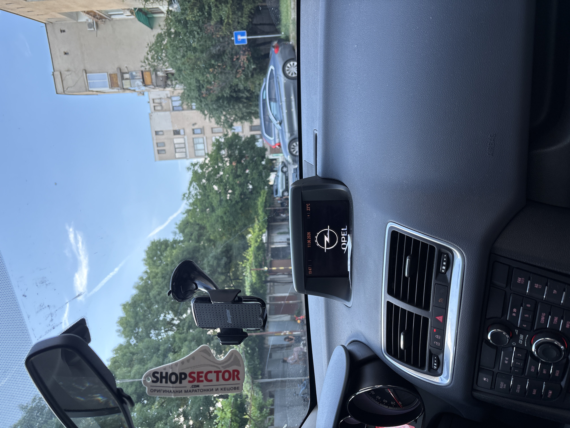 The image shows the interior of a car, viewed from the driver's perspective, with a dashboard display showing the Opel logo, the time 19:47, and the date 11.06.2026, with a temperature of 23°C. A car air freshener shaped like a shoe with the brand "SHOPSECTOR" is hanging from the rearview mirror. A phone holder is attached to the windshield. Outside the car, trees, apartment buildings, and parked cars are visible. A street sign with a blue background and a white arrow pointing down is seen on the right side. The scene suggests a sunny day in a residential area, possibly in the city of Panagyurishte, Bulgaria.